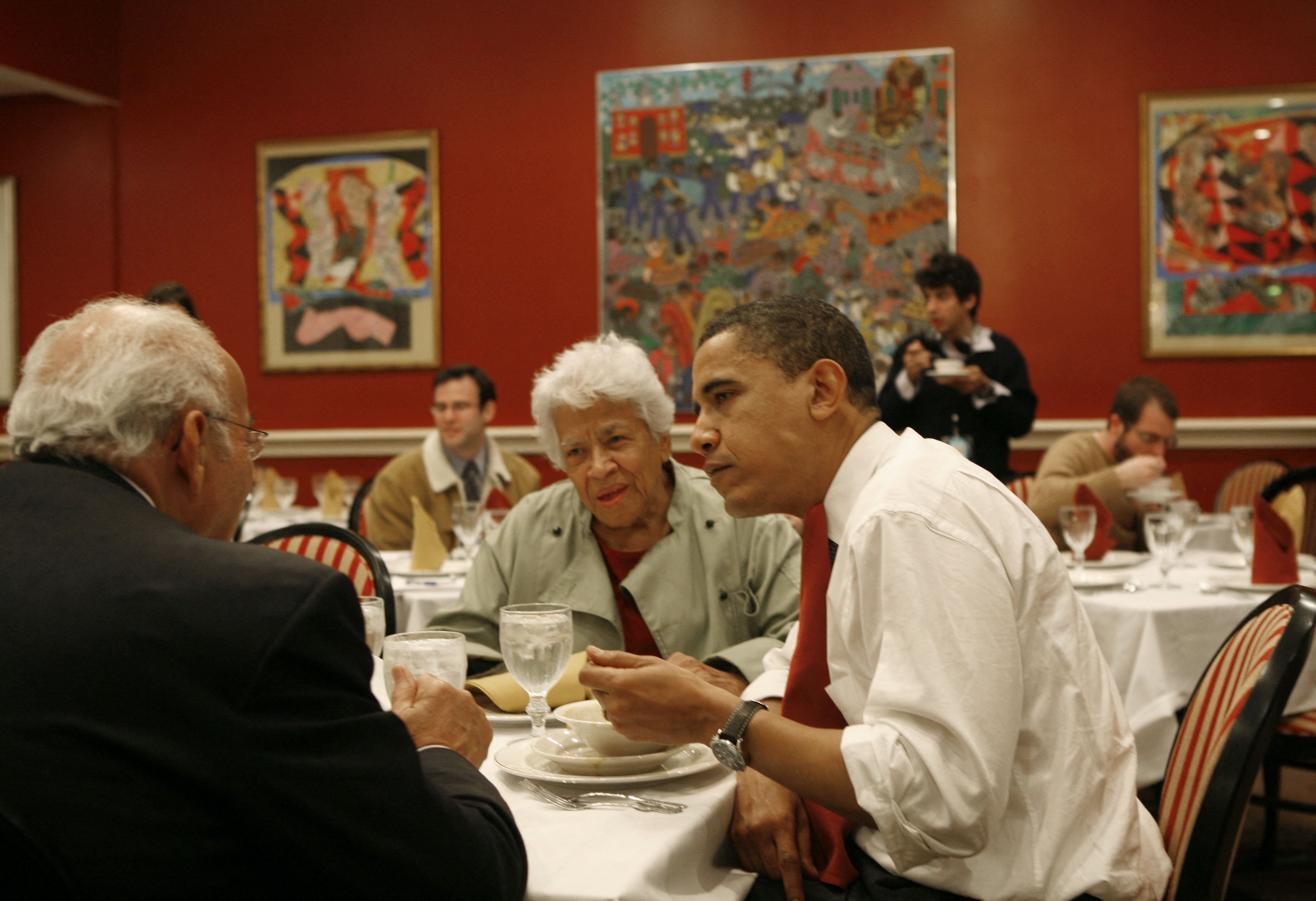 Leah Chase and Barack Obama sit around a table in a restaurant with red walls, engaged in conversation. The table is set with water glasses, silverware, and a bowl. Colorful artworks hang on the walls in the background. Other diners can be seen seated at tables throughout the room.