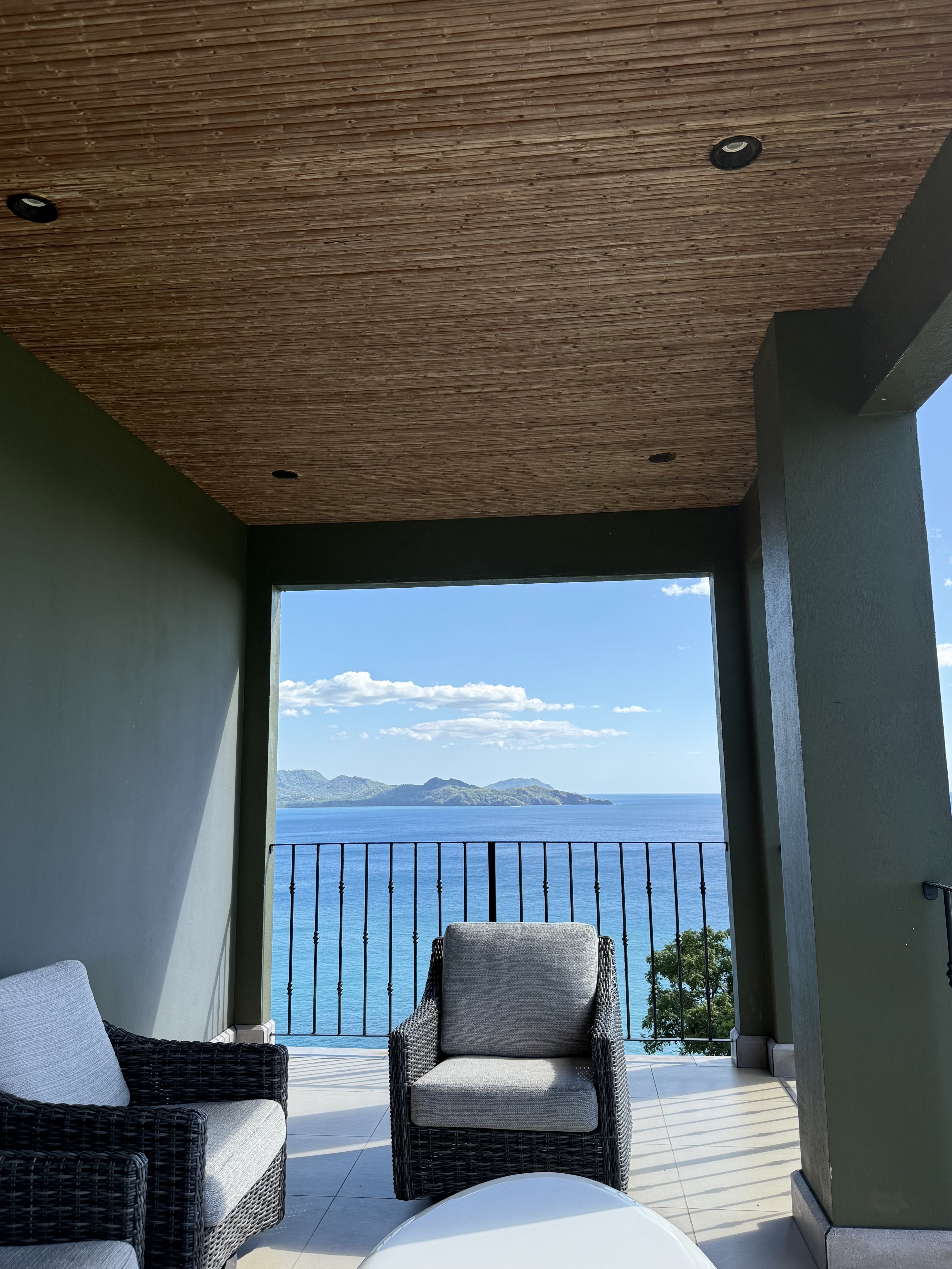 Wooden slat ceiling with recessed lights, green walls, and a balcony opening to a blue ocean and distant mountains. Wicker armchairs with light cushions face a small table, overlooking a railing.