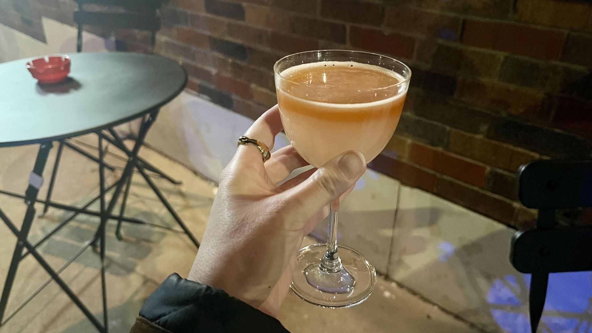 Hand holding a coupe glass with a frothy peach-colored cocktail outdoors near a brick wall, black table, and chairs at night.