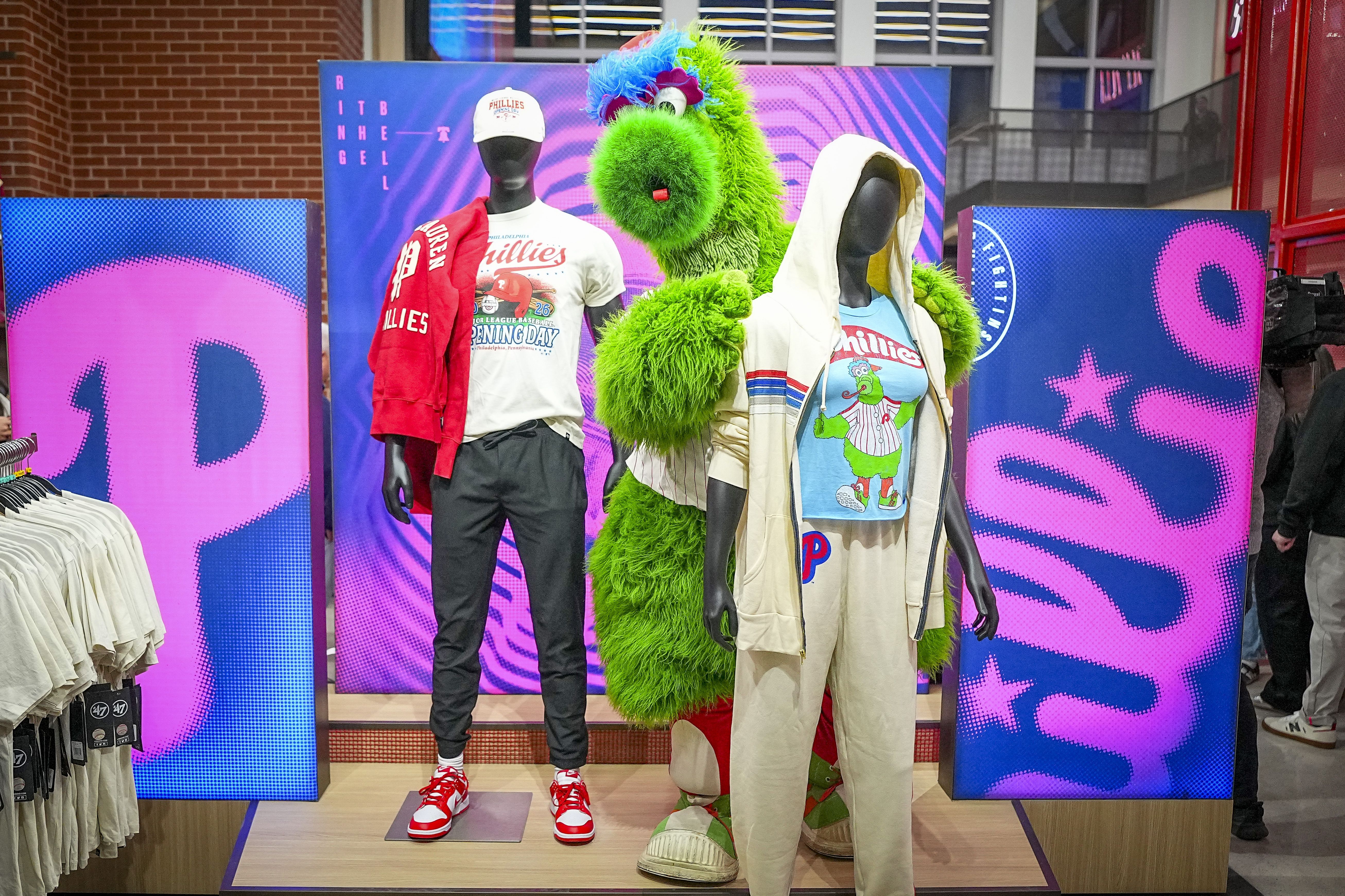 Philly-themed fashion display with two black mannequins in Phillies outfits and a bright green furry mascot behind them; pink and blue backdrops.