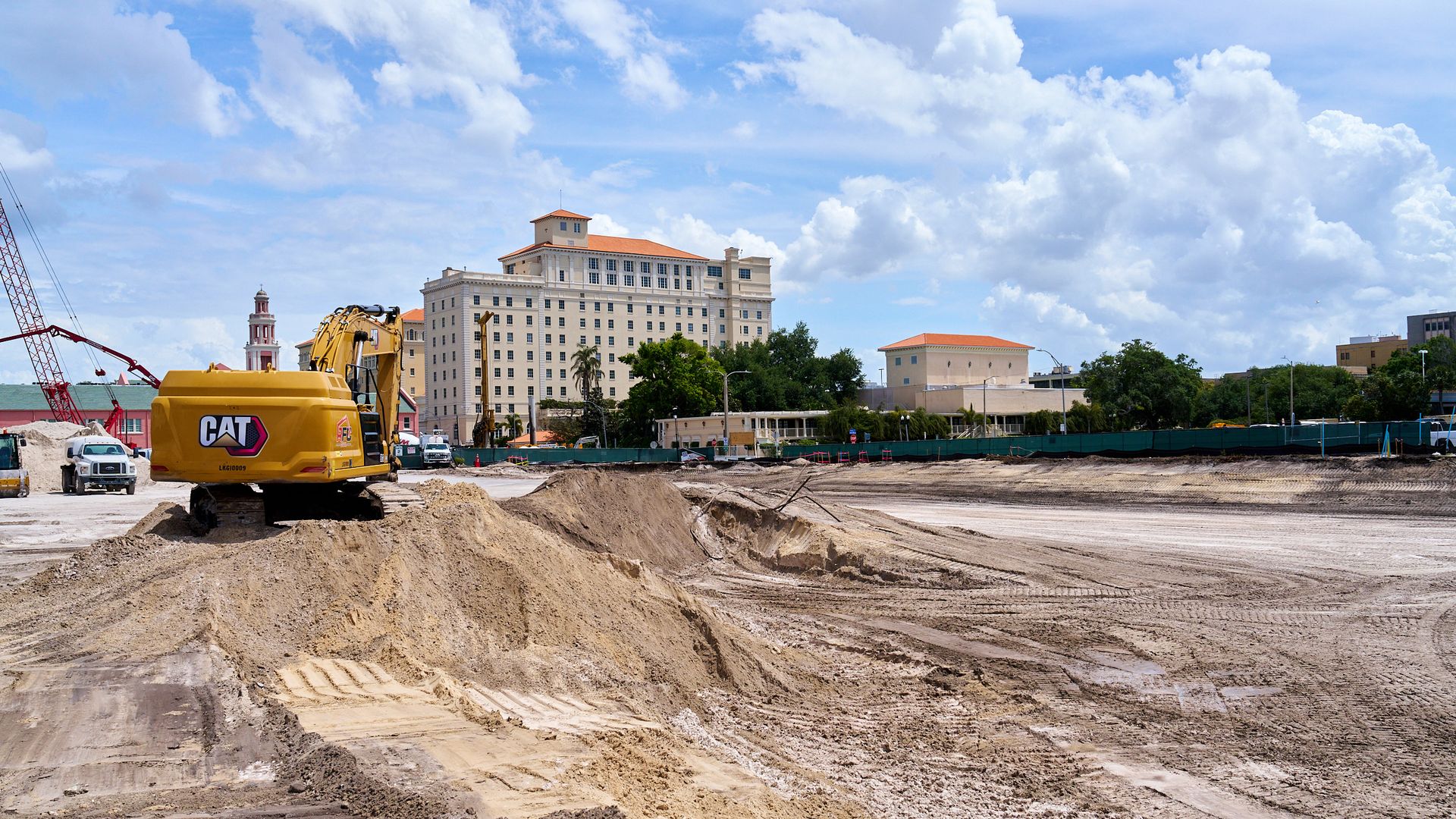 Construction site with a yellow CAT excavator in the foreground, dirt piles, a red crane on the left, and beige buildings with red-tiled roofs in the background under a bright blue sky with white clouds.