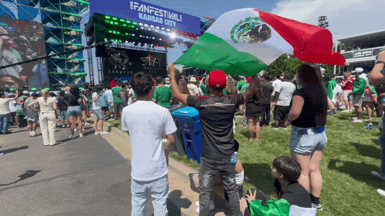Crowded outdoor festival with a large stage and screens reading "FAN FESTIVAL KANSAS CITY". In the foreground, a person waves a large Mexican flag while the crowd watches on grass and pavement.