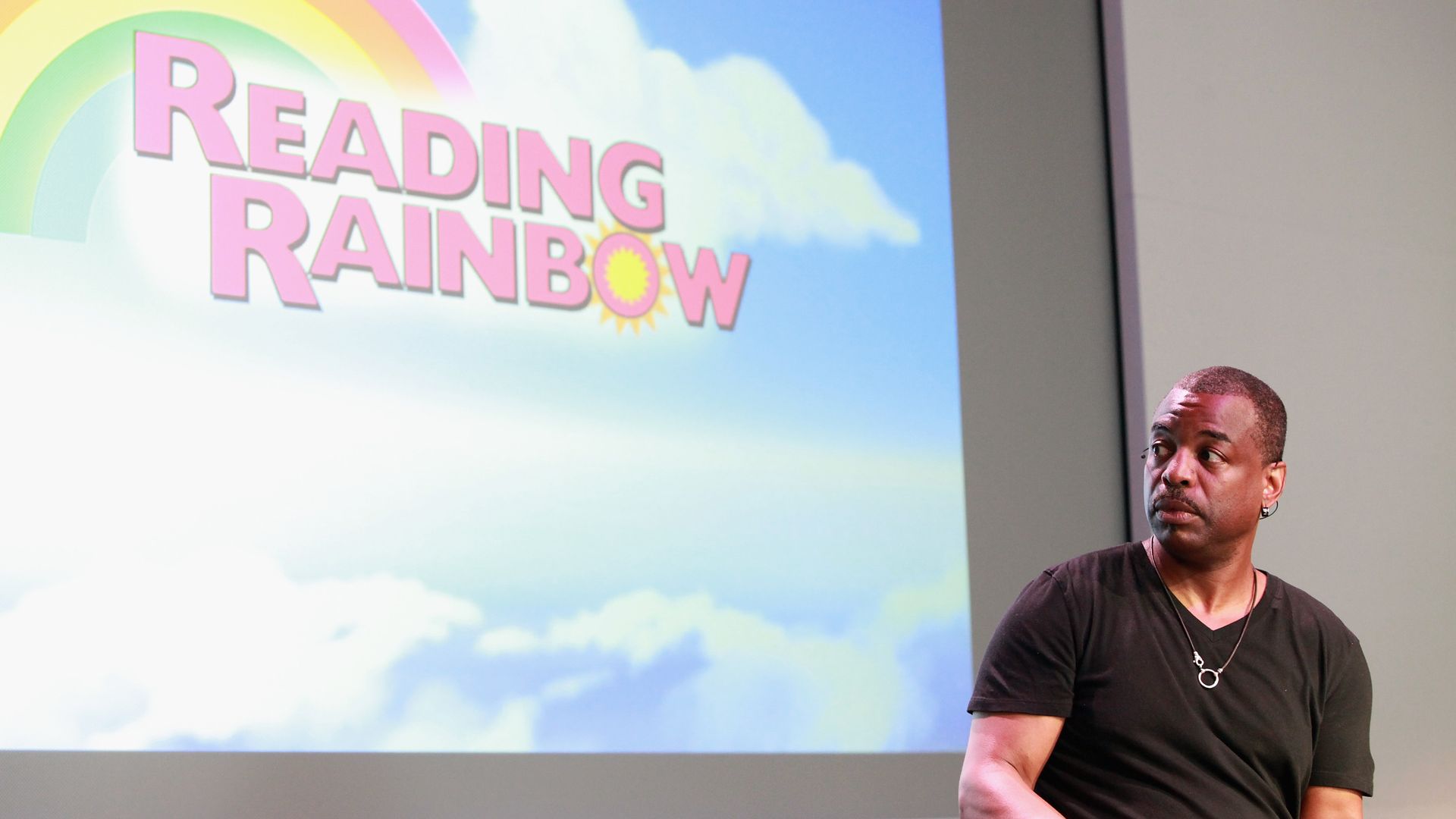 LaVar Burton sitting and looking at a screen that has the "Reading Rainbow" logo on it.