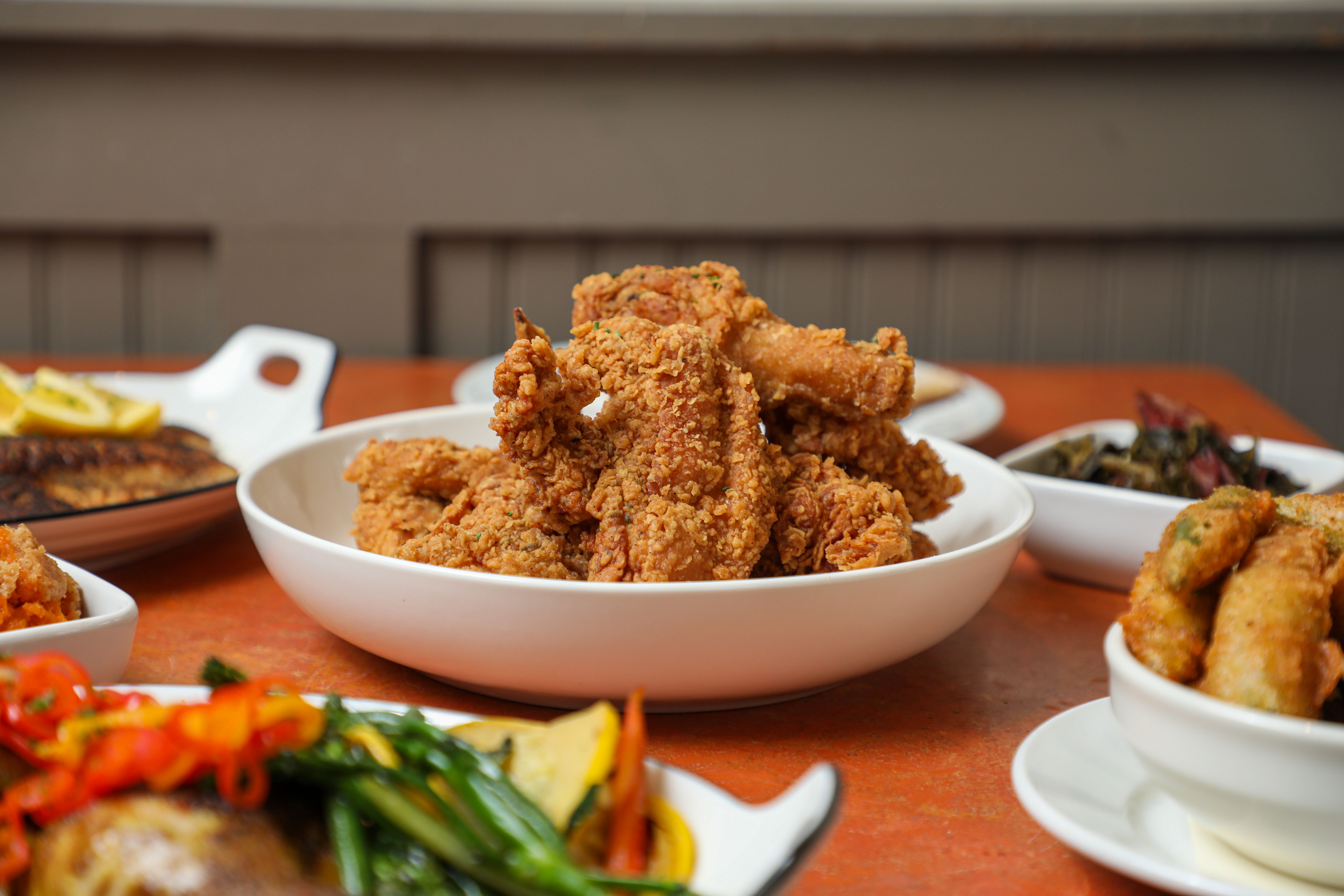 An orange table top is filled with white plates and bowls filled with Creole soul food staples, like fried fish, potatoes, green beans and fried catfish.