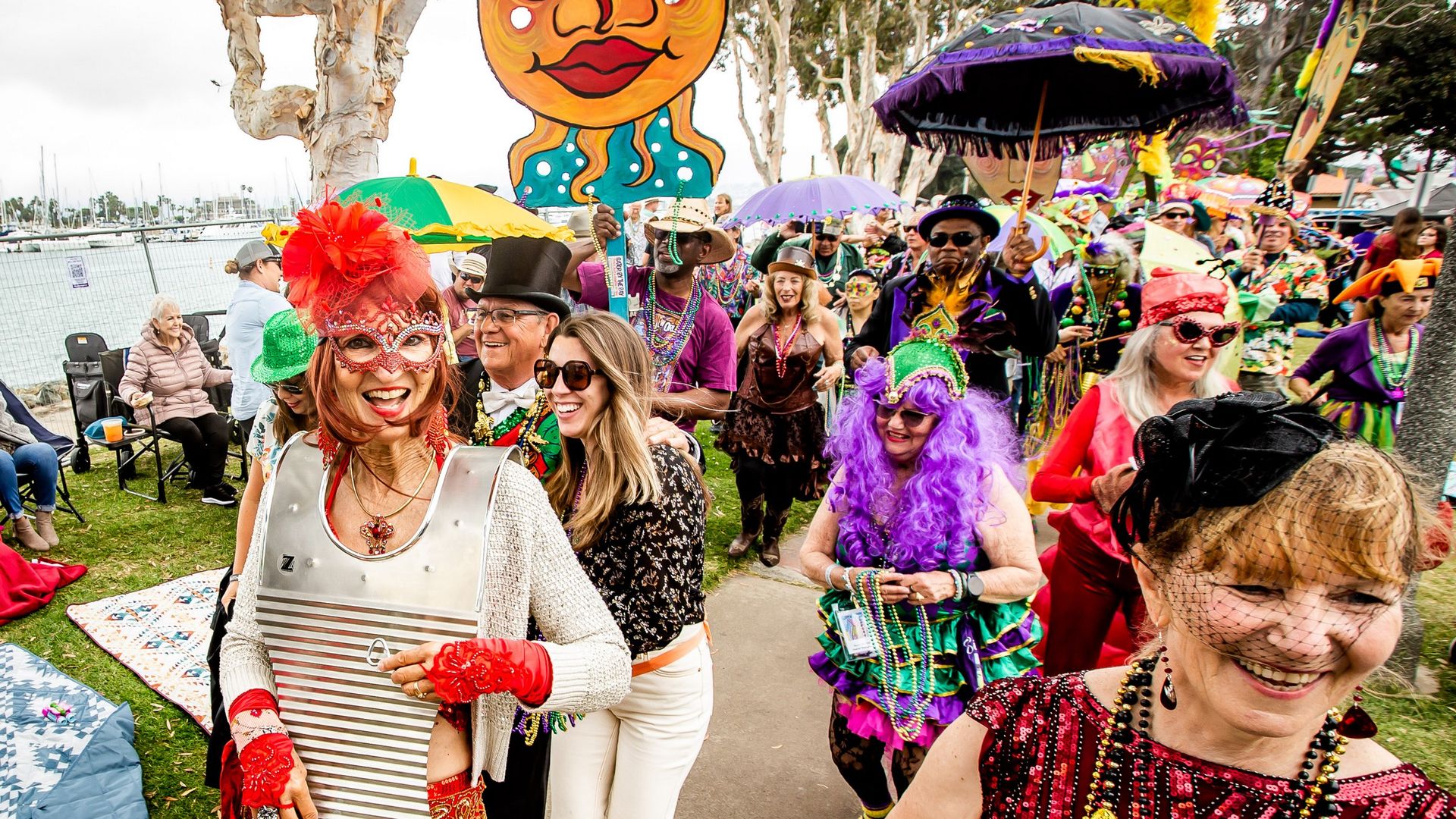 People dressed in festive Mardi Gras attire walk in a parade by the bay.