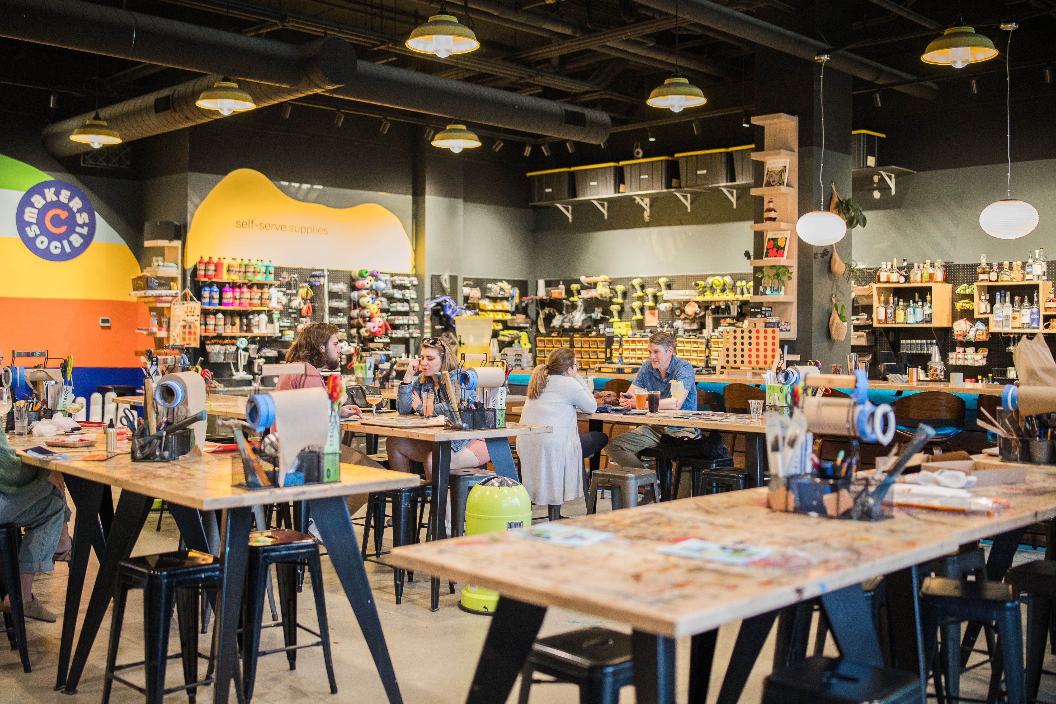 Creative workspace with wooden tables, art supplies, and stools. People are seated, chatting. Shelves stocked with tools and colorful supplies. Warm, industrial lighting overhead.