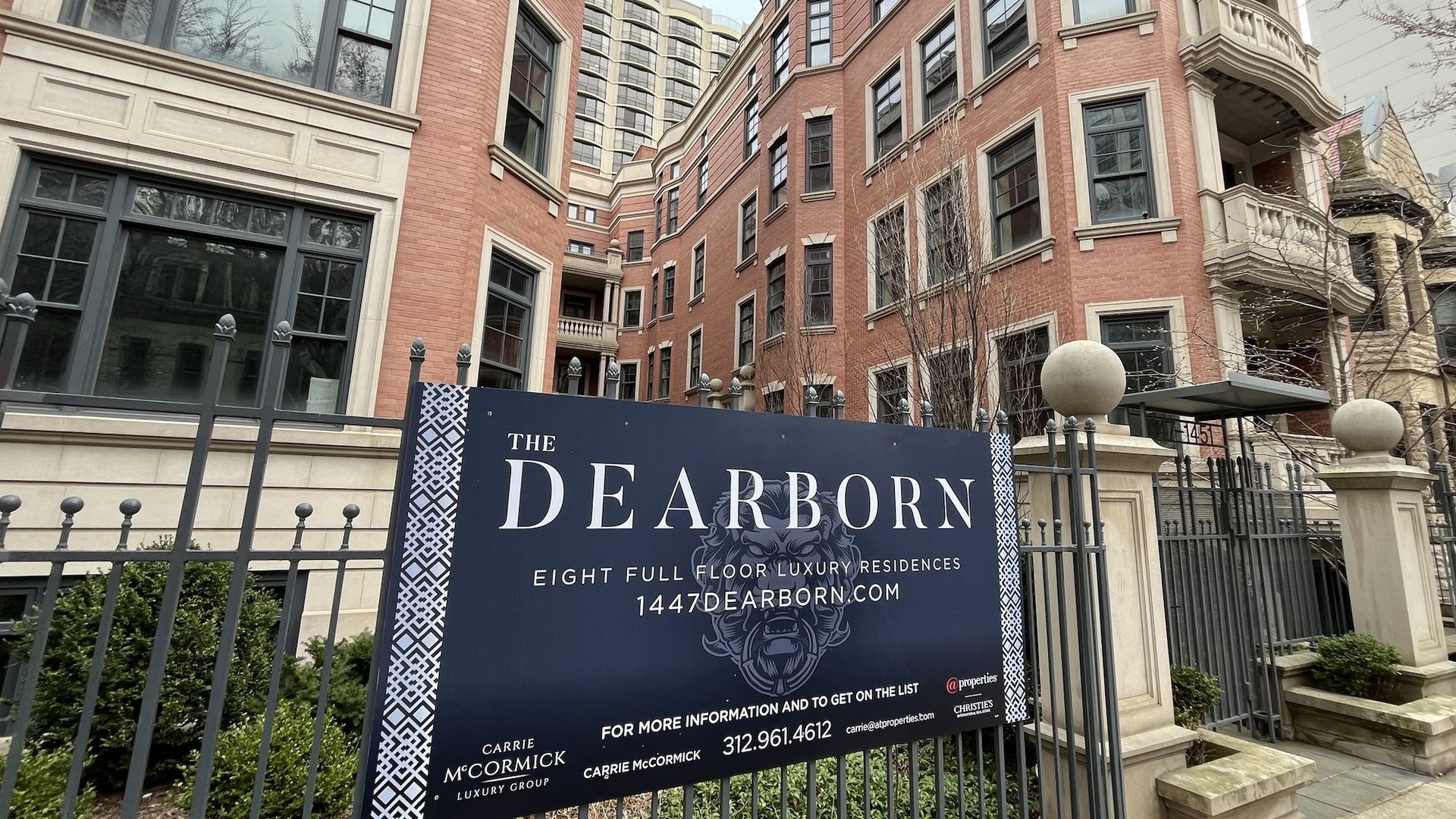 Brick residential complex with large windows. A navy sign reads "The Dearborn" advertising eight full-floor luxury residences. Iron fence, stone spheres, and curved balconies with a high-rise behind.