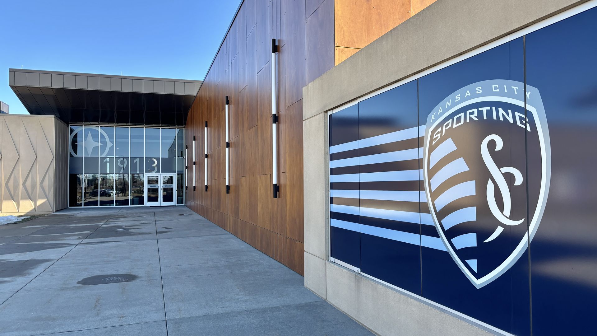 Exterior of Sporting Kansas City building with blue logo on right, brown wood paneling on wall, and glass doors displaying star and "1913" under clear blue sky.