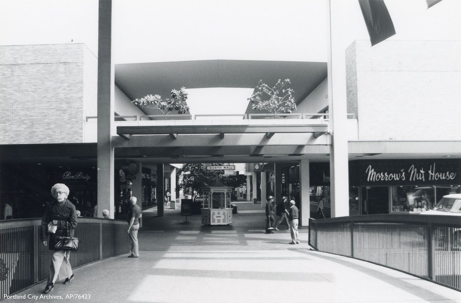 Black-and-white photo of an outdoor shopping mall corridor with a small kiosk in the center, people walking and standing, stores including "Morrow's Nut House" visible on the right.