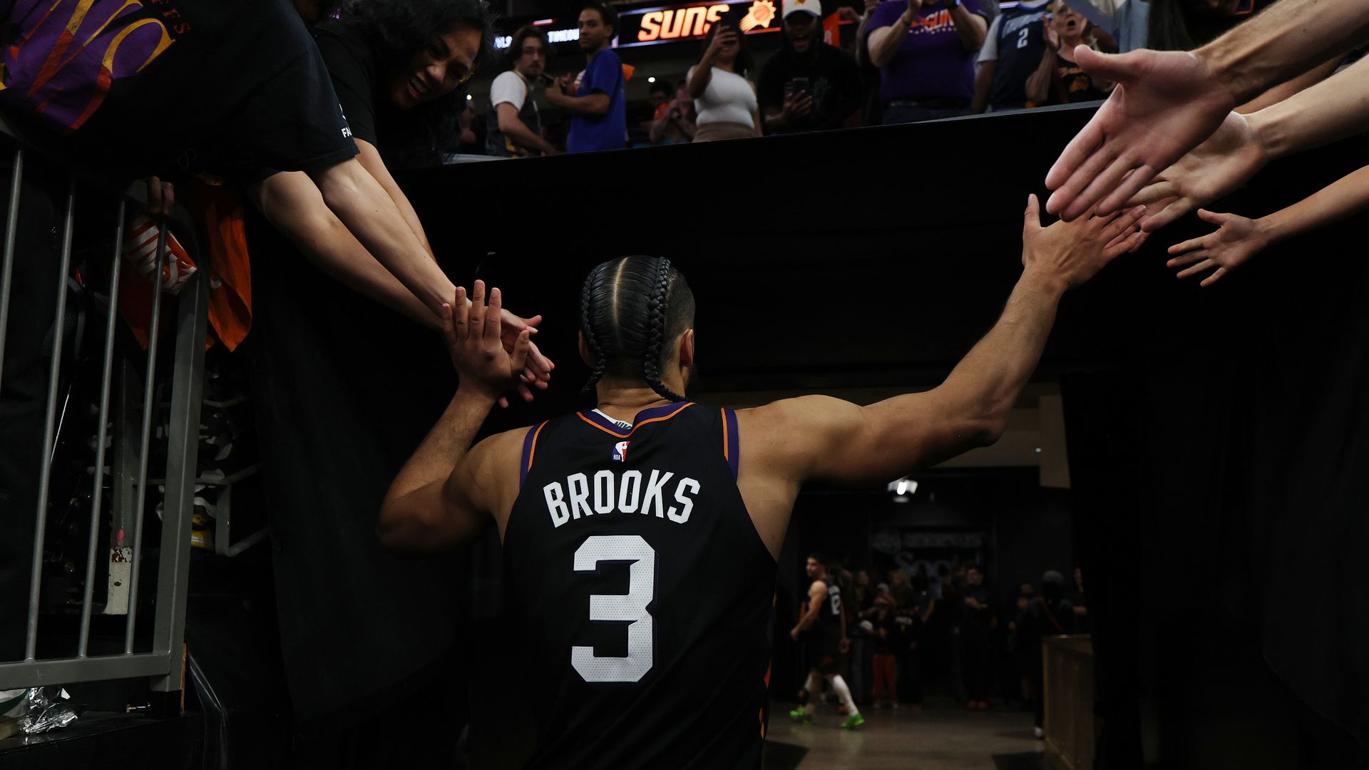 A basketball player in a black jersey, viewed from behind, with the word "Brooks" and the number "3" on the back of his uniform, slaps hands with fans on both sides of him as he walks off the court and into the tunnel. 