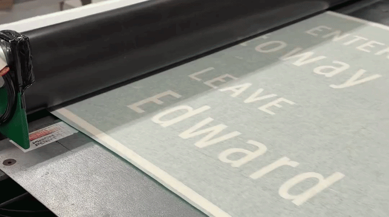 Close-up of a large-format printer feeding a sheet that reads "DON'T LEAVE EDWARD" in large white letters on a pale gray background; printer head visible on the left.