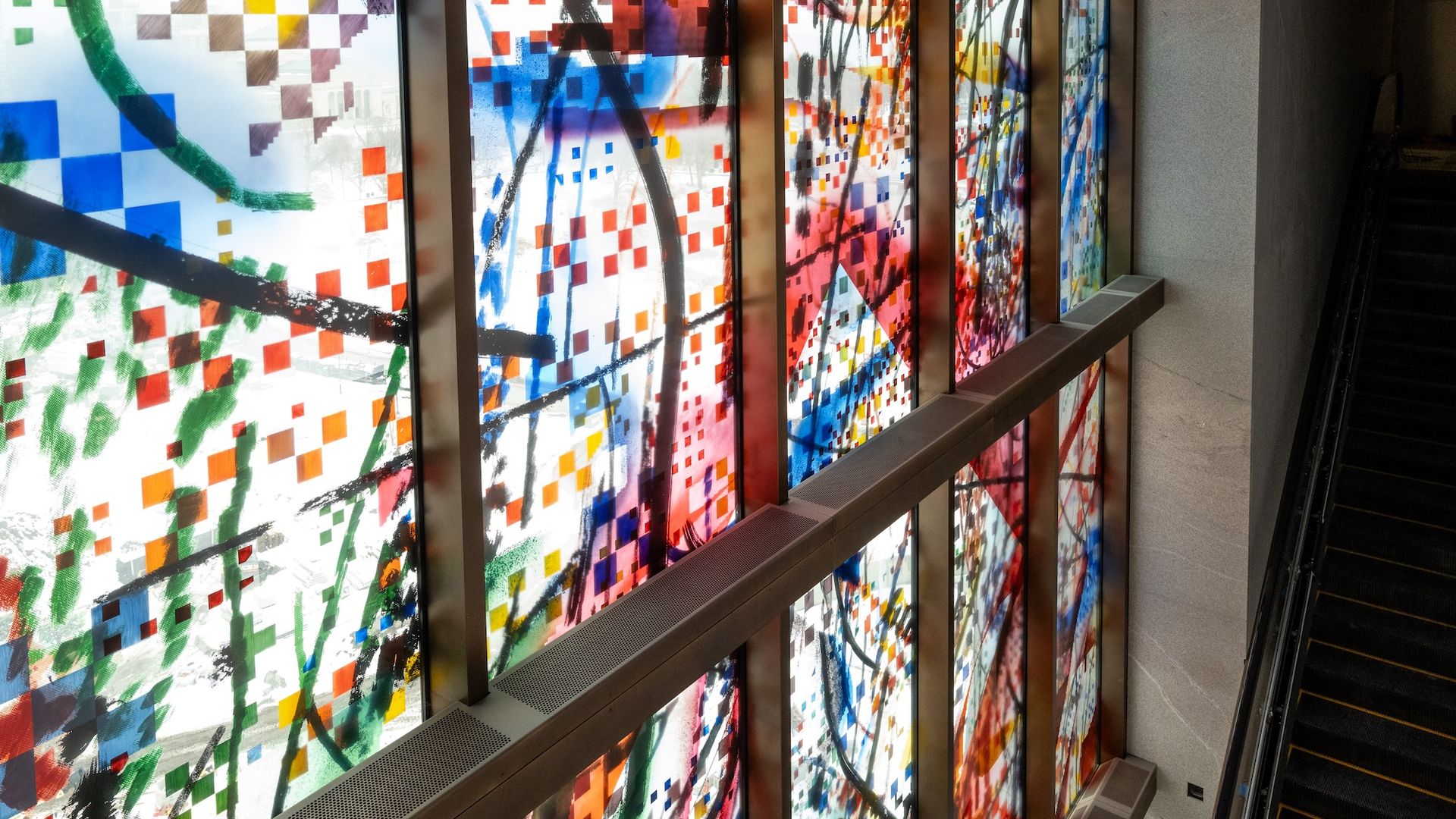 Colorful abstract stained-glass panels line a stairwell wall, filled with small multicolored squares and sweeping black brush strokes; a metal handrail runs along the glass beside a dark staircase.
