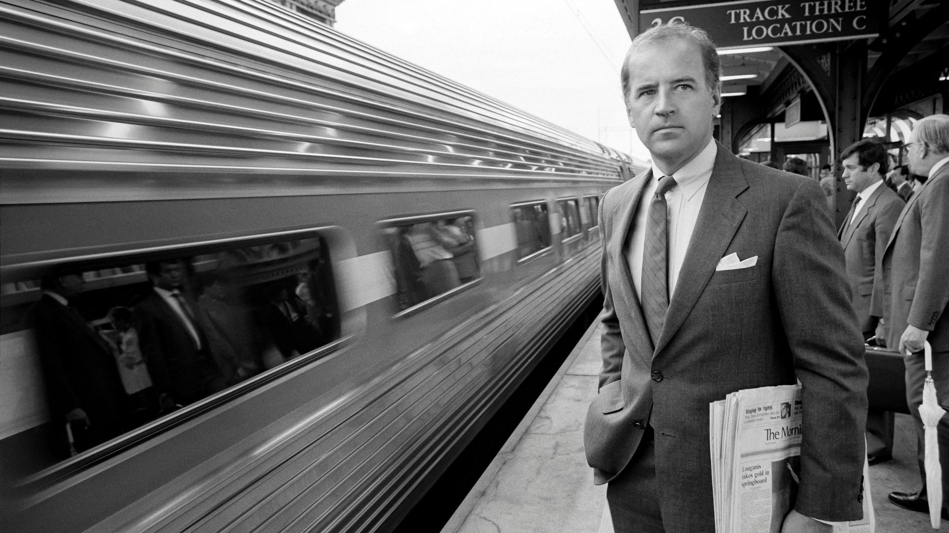 Biden holds a newspaper in a black and white photograph while waiting for a train