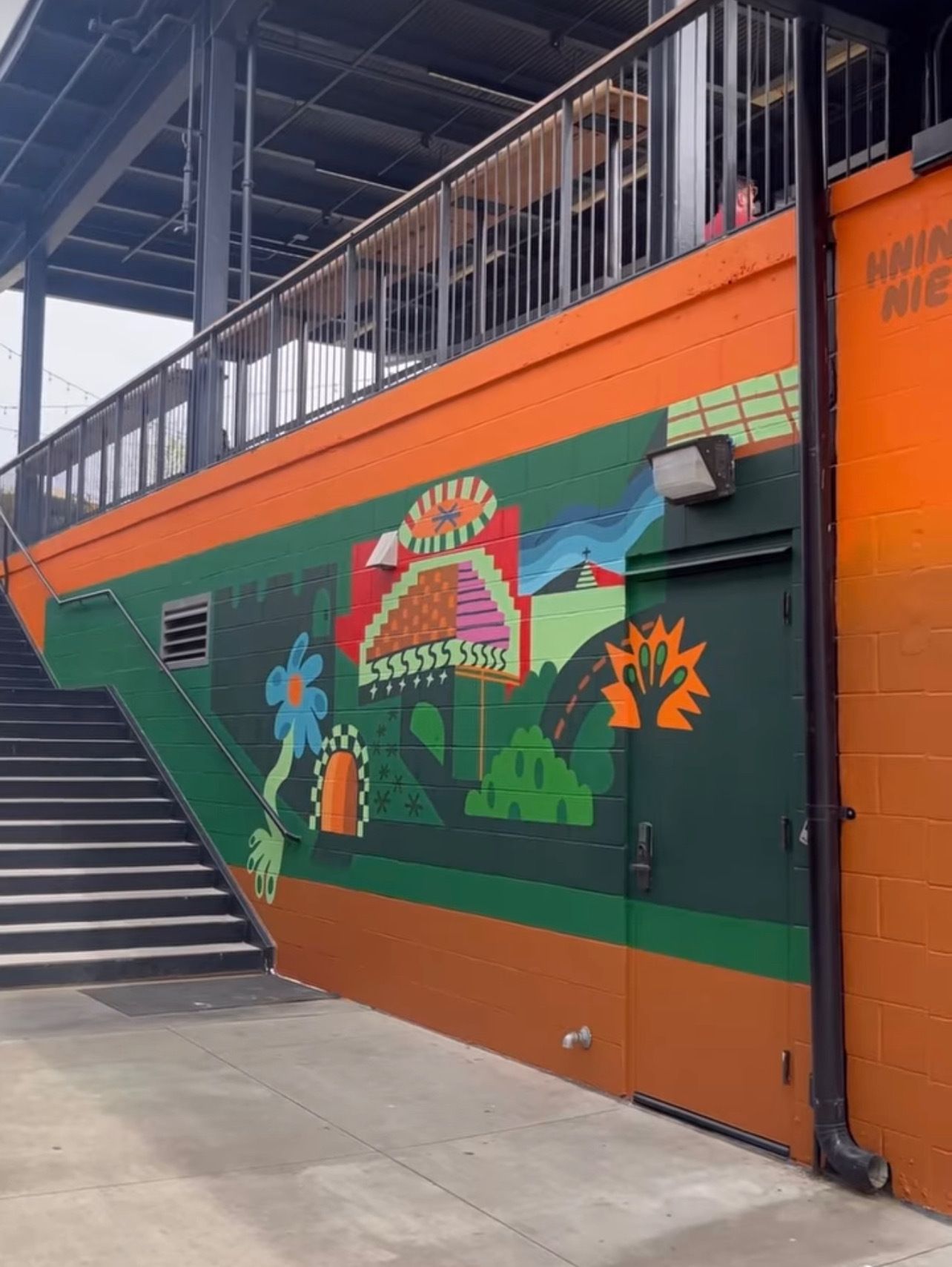 Outdoor wall under a walkway with an orange border and green mural. A colorful central pyramid and abstract shapes sit among hills, with a left stairway, railing, a door, and a right downspout.