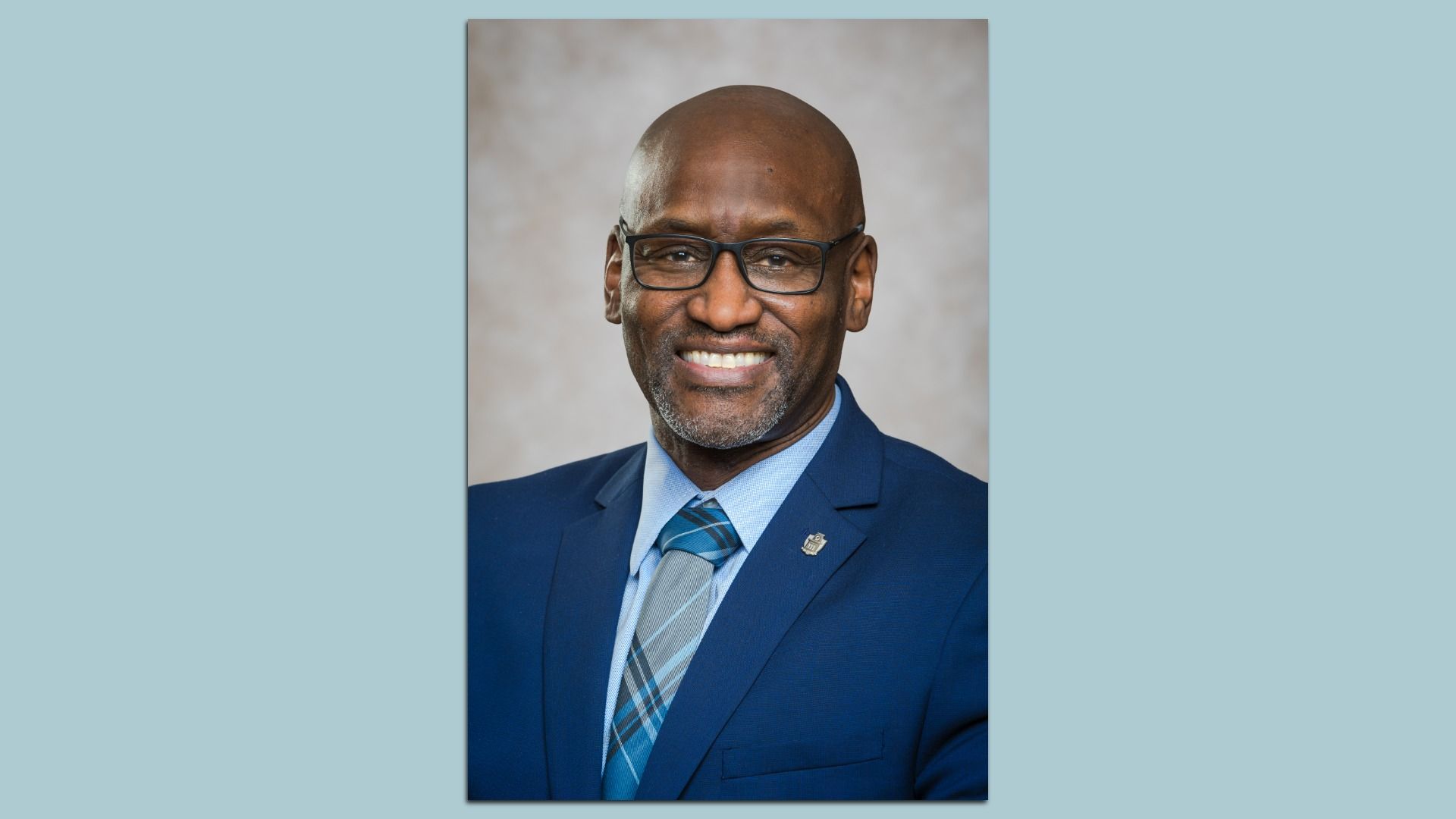 A smiling man with glasses and a bald head wears a blue suit jacket, light blue shirt, and a blue plaid tie. He stands in front of a soft, neutral-colored backdrop for a formal portrait.