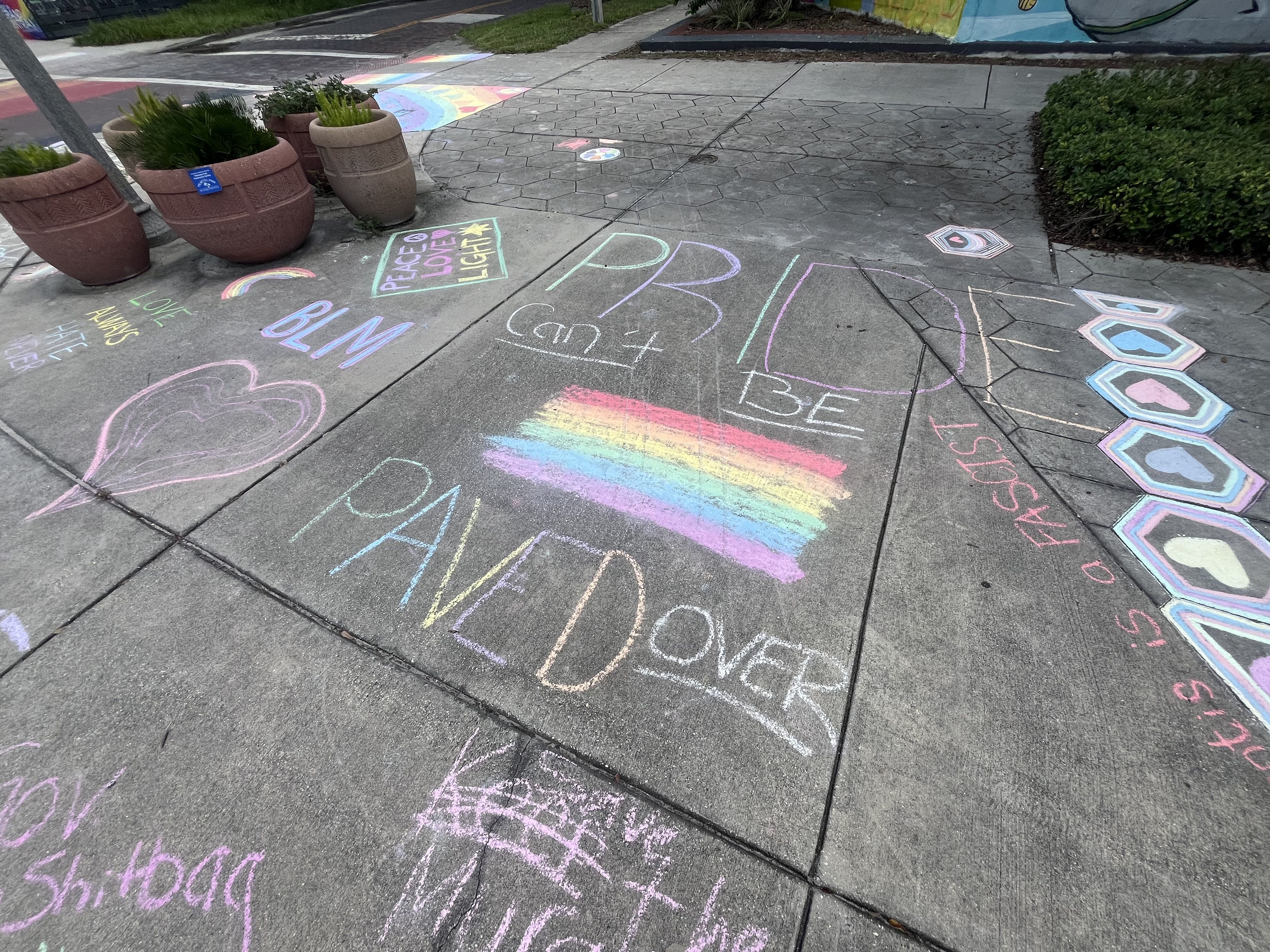 Colorful sidewalk chalk art with messages including a large rainbow and "PRIDE can't BE PAVED OVER," "BLM," a pink heart, rainbows, and phrases like "PEACE LOVE LIGHT" and "LOVE ALWAYS WINS."