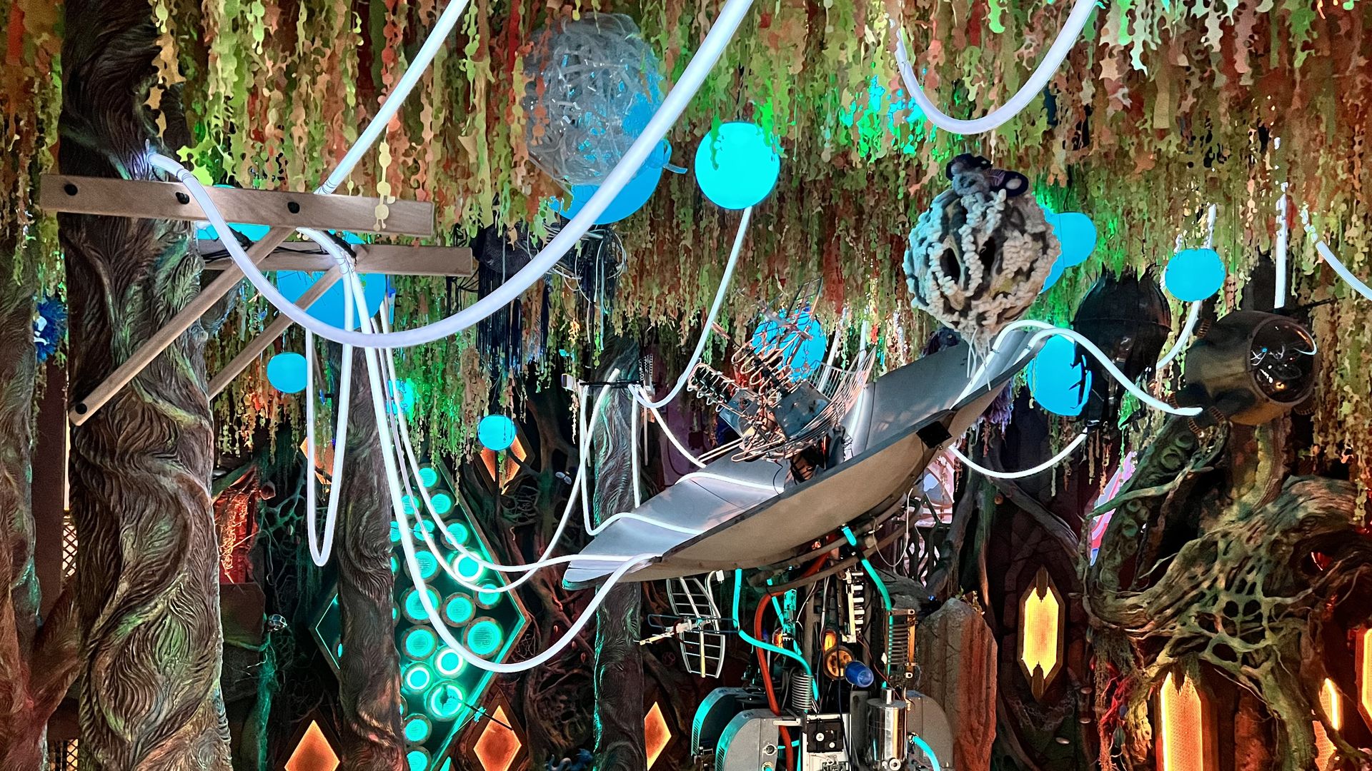 Futuristic, dimly lit room with tree-like pillars, neon blue and orange lights, hanging tubes, glowing blue spheres, and a suspended chair surrounded by mechanical and electronic components.