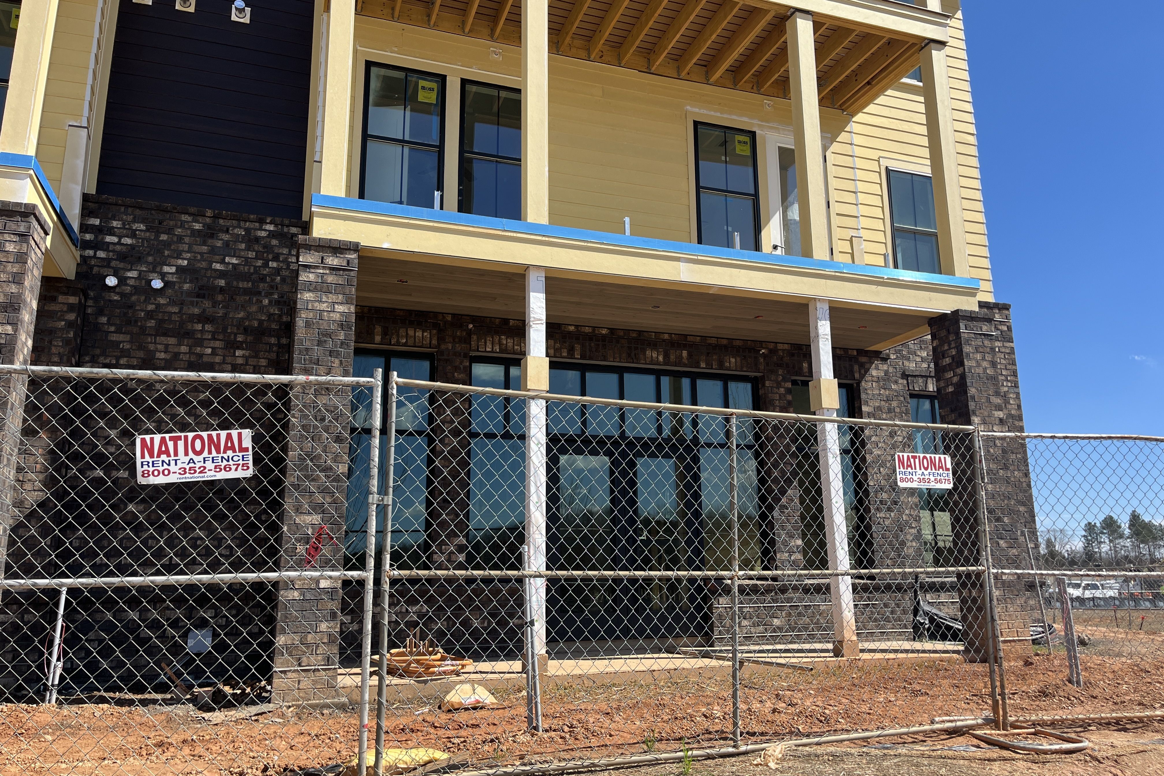 Under-construction, two-story building with brick base and yellow siding, large glass doors and windows, brick columns, chain-link fence with National Rent-A-Fence signs, dirt ground, blue sky.
