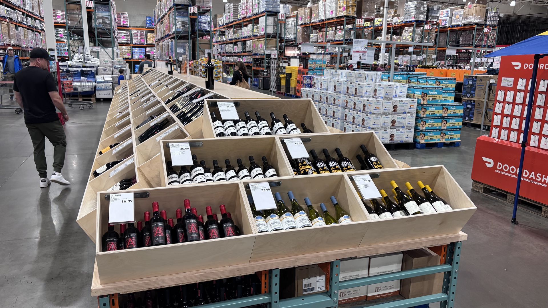 Inside a warehouse-style store, a wooden wine display dominates the foreground with bottles. A man in a black shirt and cap stands left; tall shelves and pallets with baby wipes visible in the background.