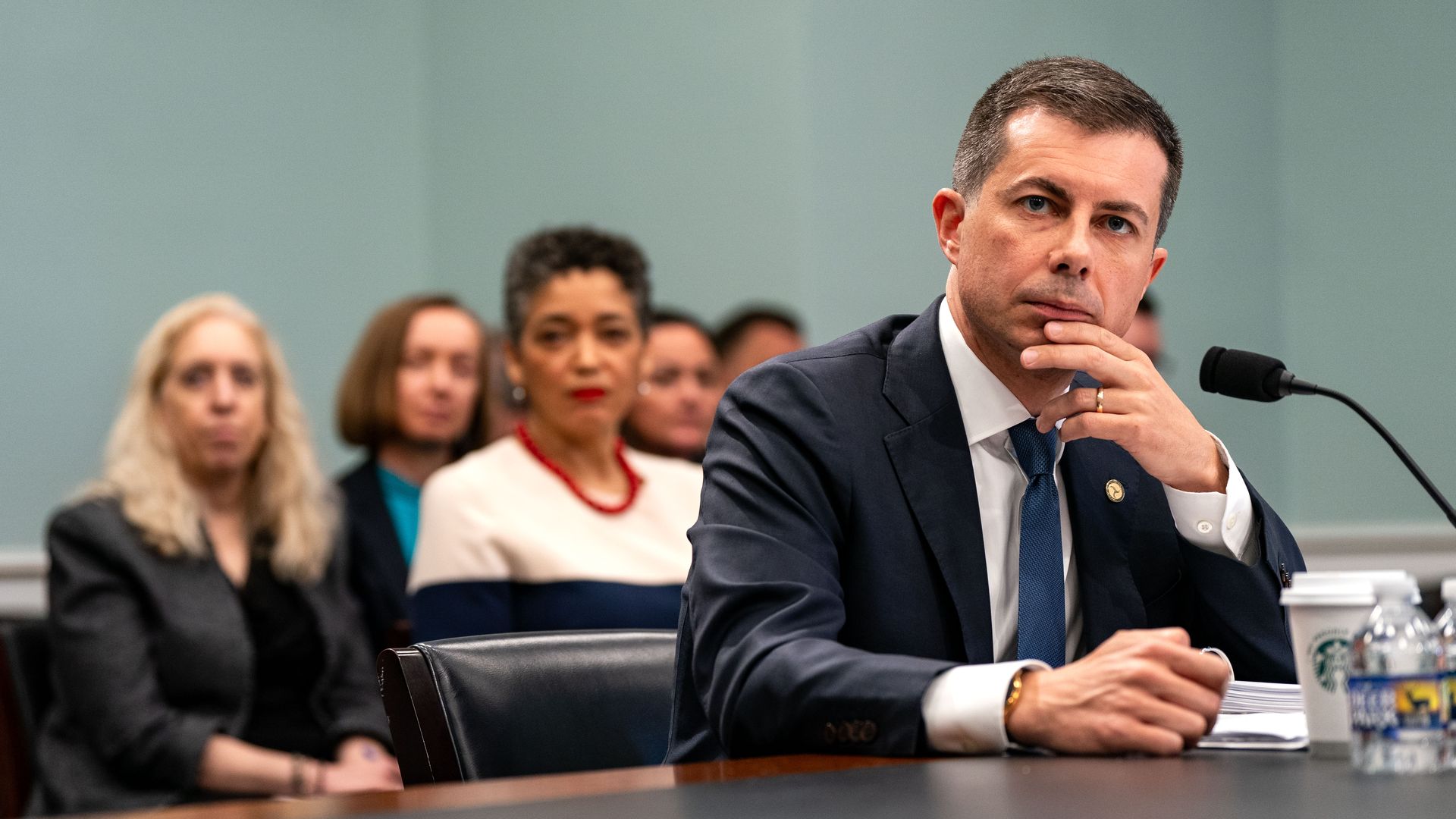 Former Secretary of Transportation Pete Buttigieg, who has black hair that's peppered with gray and who is wearing a navy jacket with a gold pin, a white shirt and dark blue tie, puts his hands to his mouth as he stairs ahead at a mic on a desk while testifying at the U.S. Capitol.