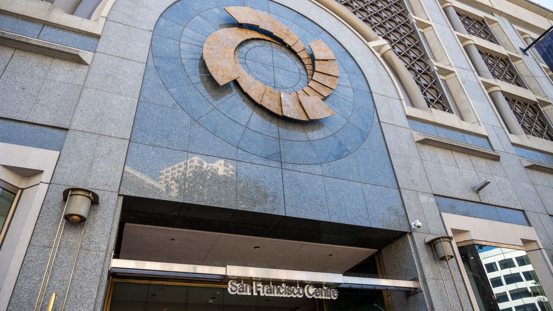 Facade of the San Francisco Centre: a large rust-colored circular metal sculpture set in a blue circular stone panel above the entrance, flanked by gray stone walls and grid-pattern windows.