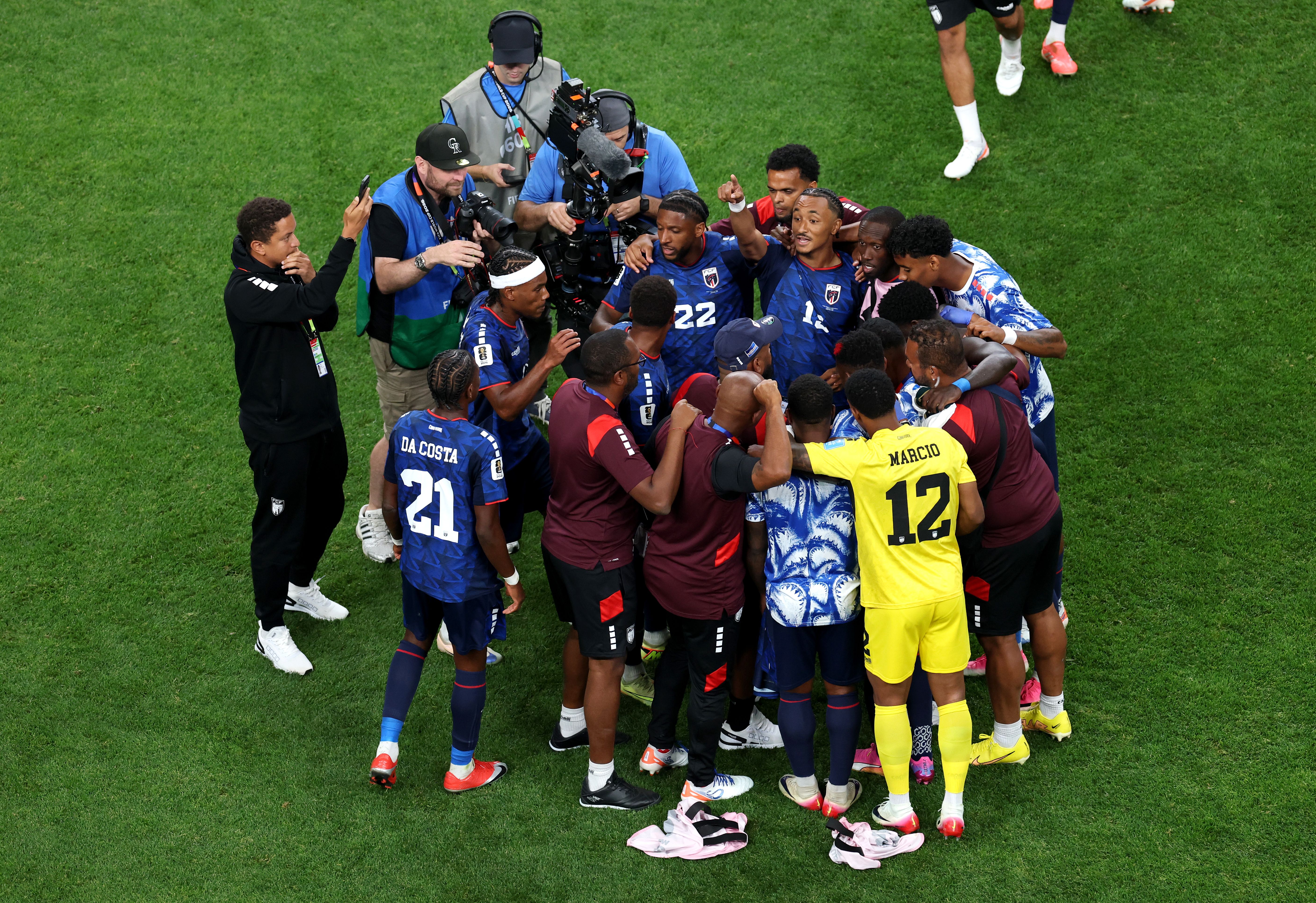 Soccer players in blue uniforms form a tight huddle on a green field, with a yellow-jersey goalkeeper nearby; photographers in blue vests circle, filming the moment.