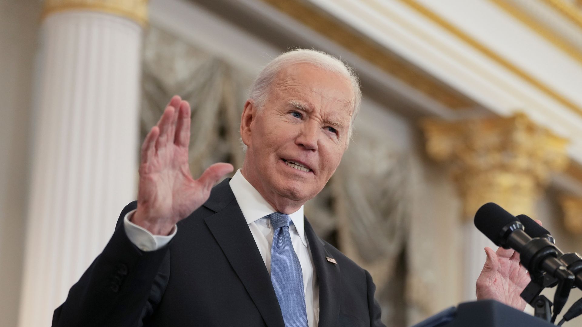 President Biden holds up his right hand while speaking into a microphone