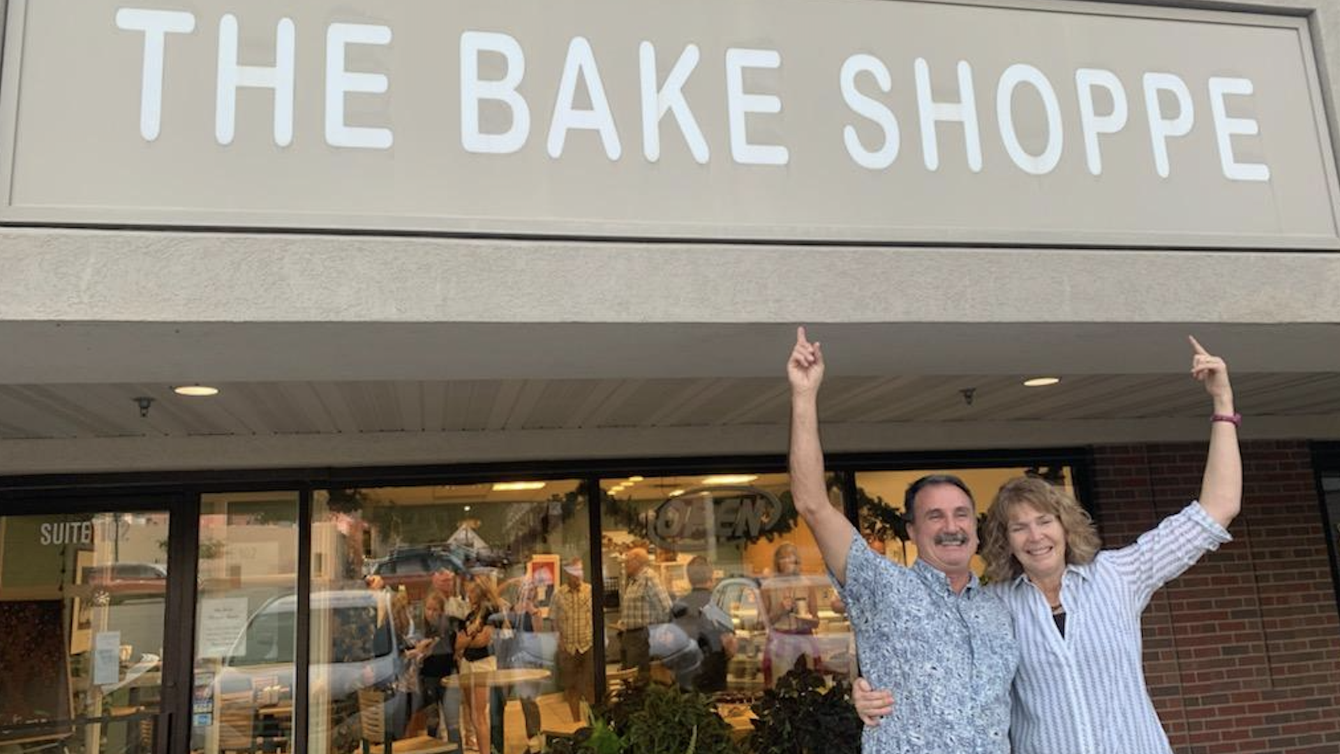 Two smiling people outside a bakery raise their arms under a large sign reading "THE BAKE SHOPPE." The man wears a blue patterned shirt, the woman a striped blouse; warm lights glow inside.