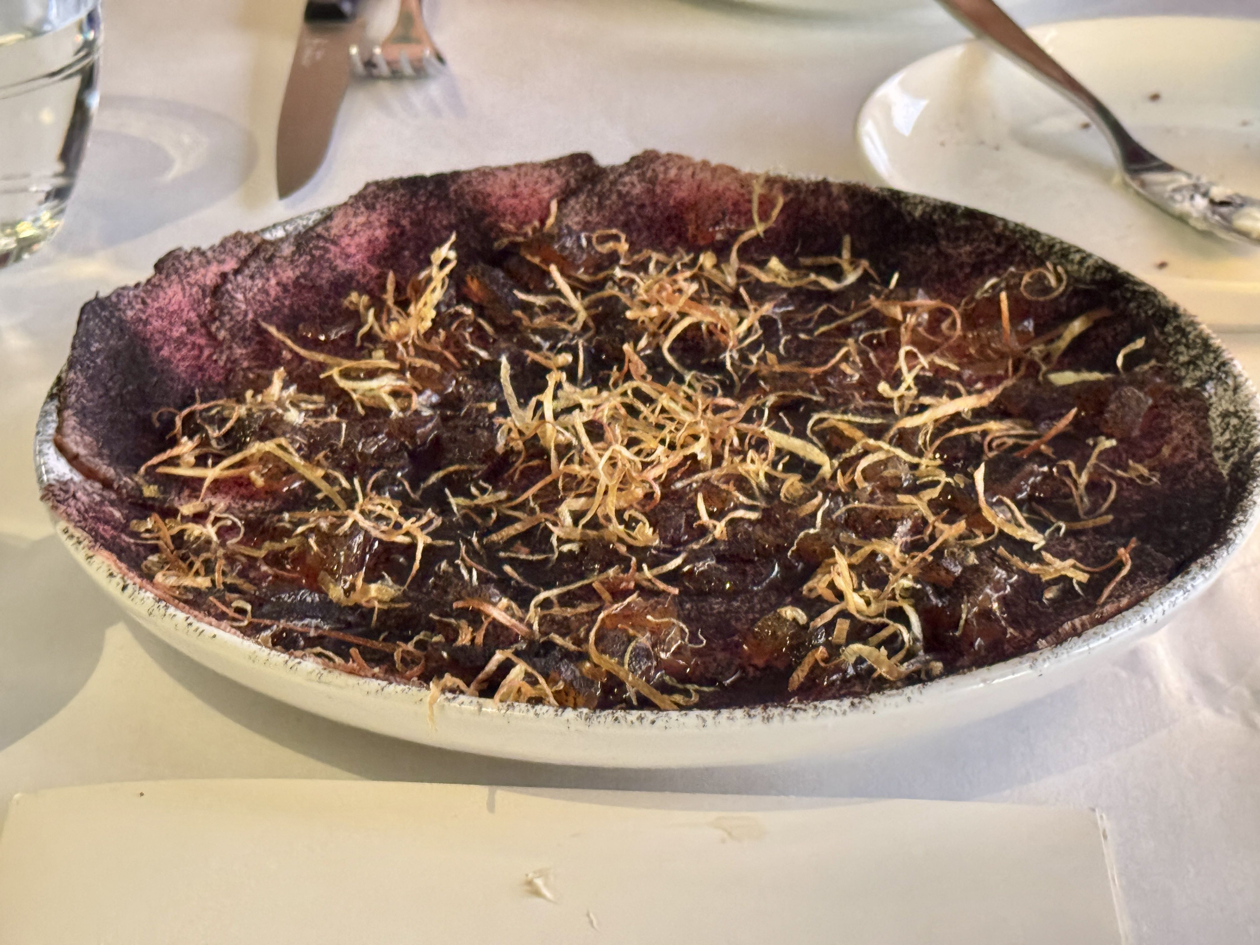 Plate with a dark purple, cheesecake-like dessert topped with golden brown crispy strands, on a white table with a knife, fork, and empty glass in the background.