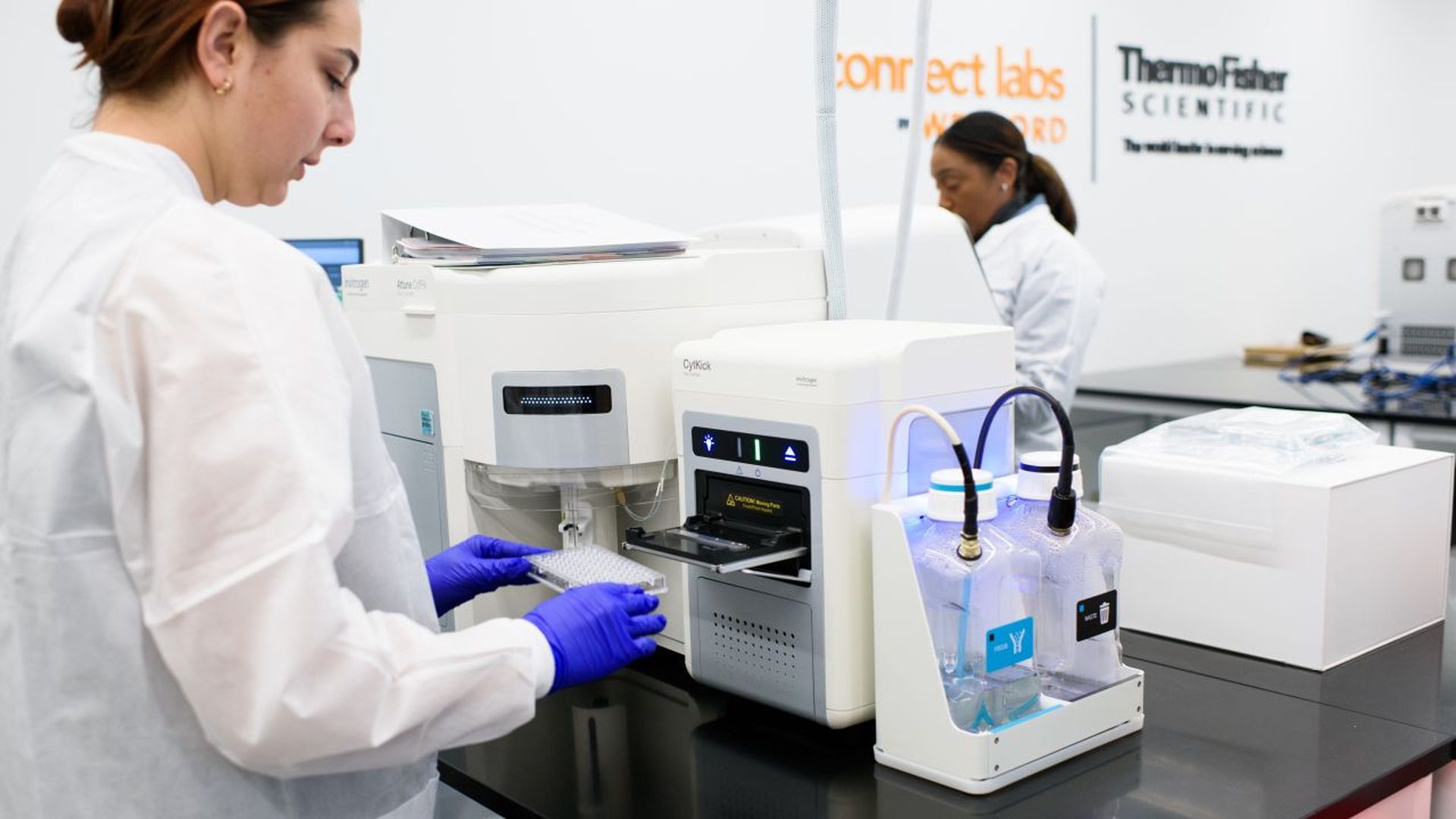 Lab scene with a technician in a white coat and purple gloves operating a white analytical instrument on a black bench; another researcher works in the background, with Thermo Fisher Scientific signage on the wall.