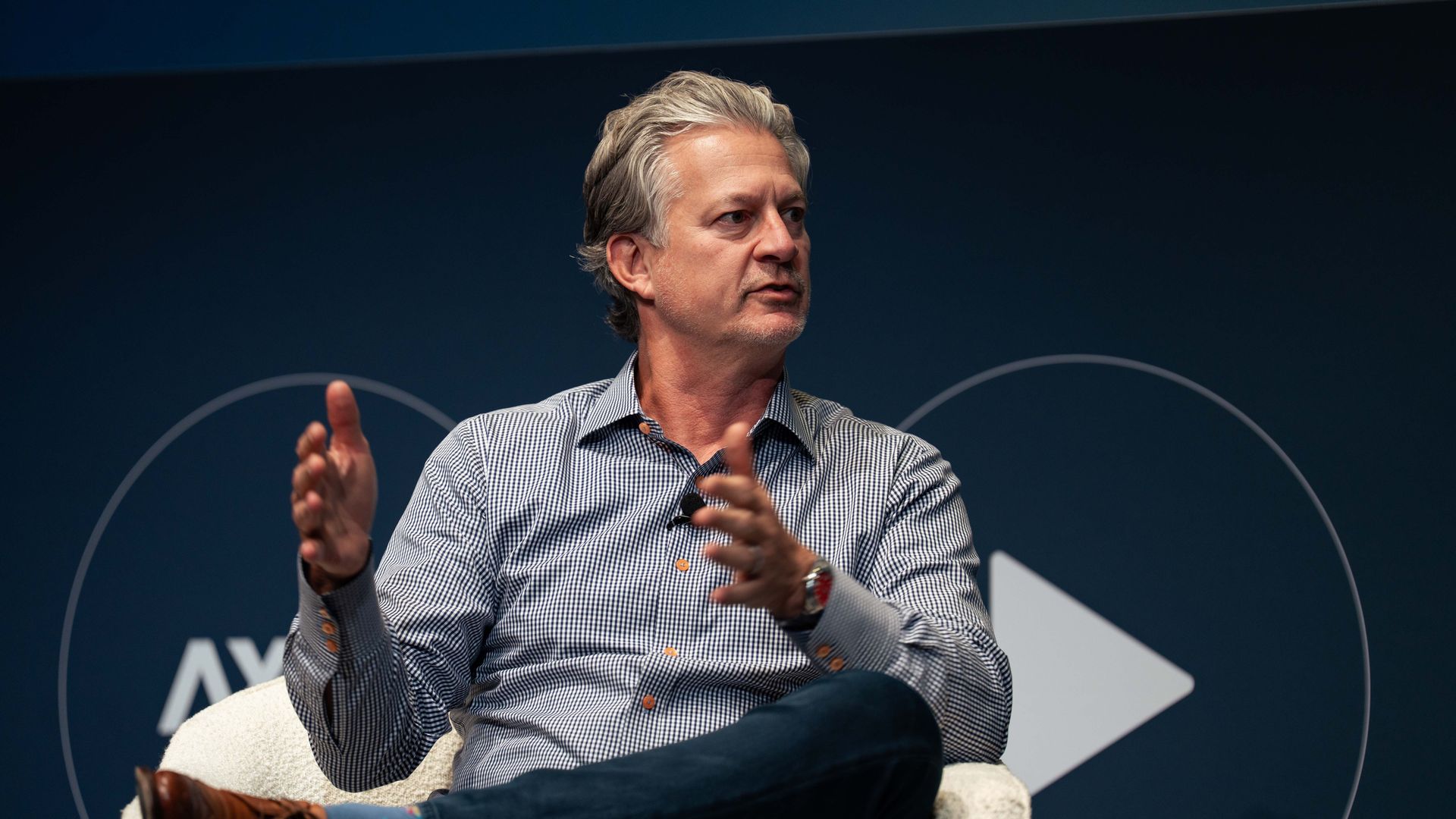 A gray-haired man in a checked shirt sits in a chair on a dark blue stage, gesturing with his hands while speaking. He wears jeans and brown shoes, with faint white curved lines in the background.