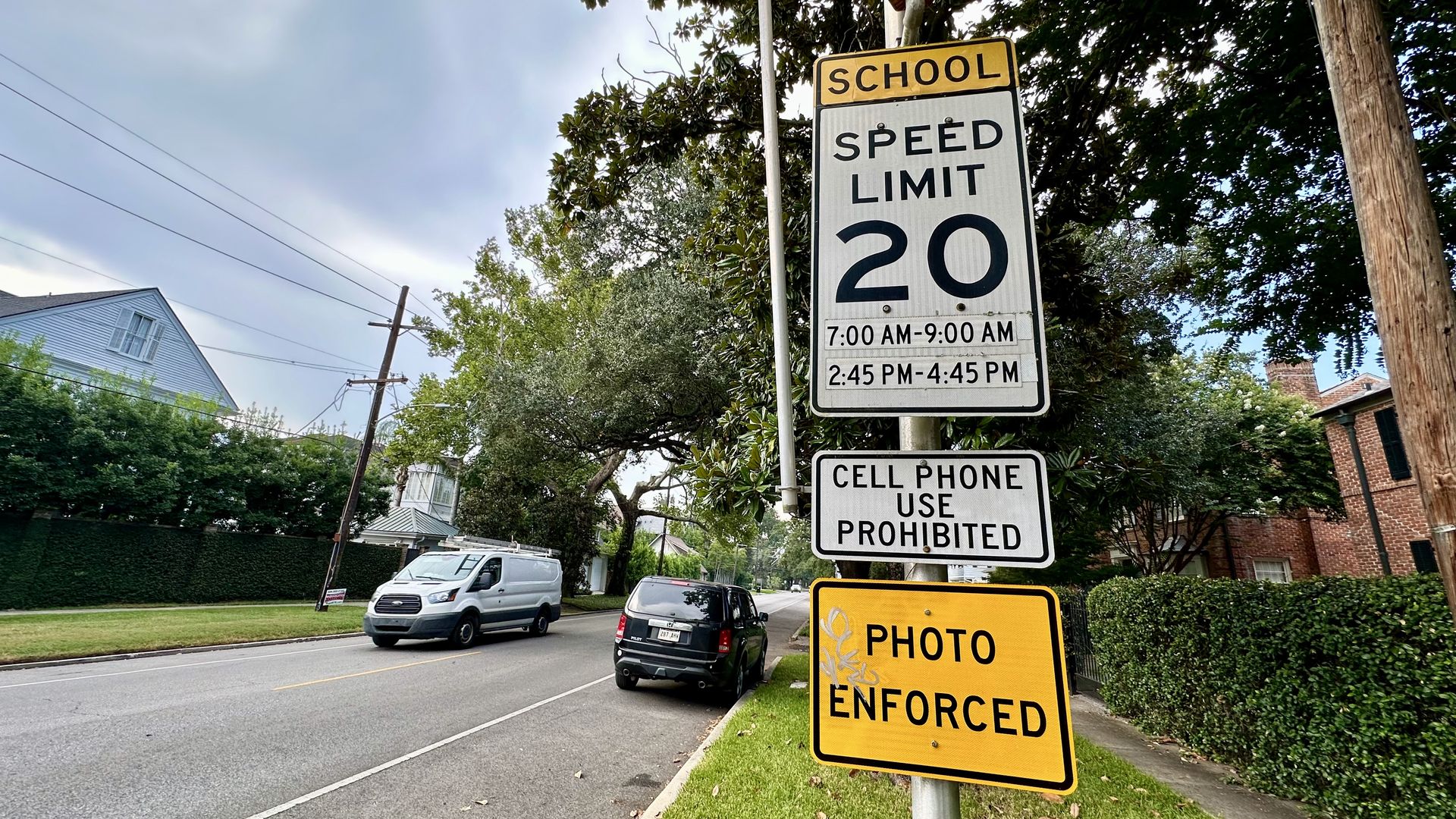 Street with school zone signs showing speed limit 20 mph during specific morning and afternoon hours, cell phone use prohibited, and photo enforcement in effect, near parked cars and houses.
