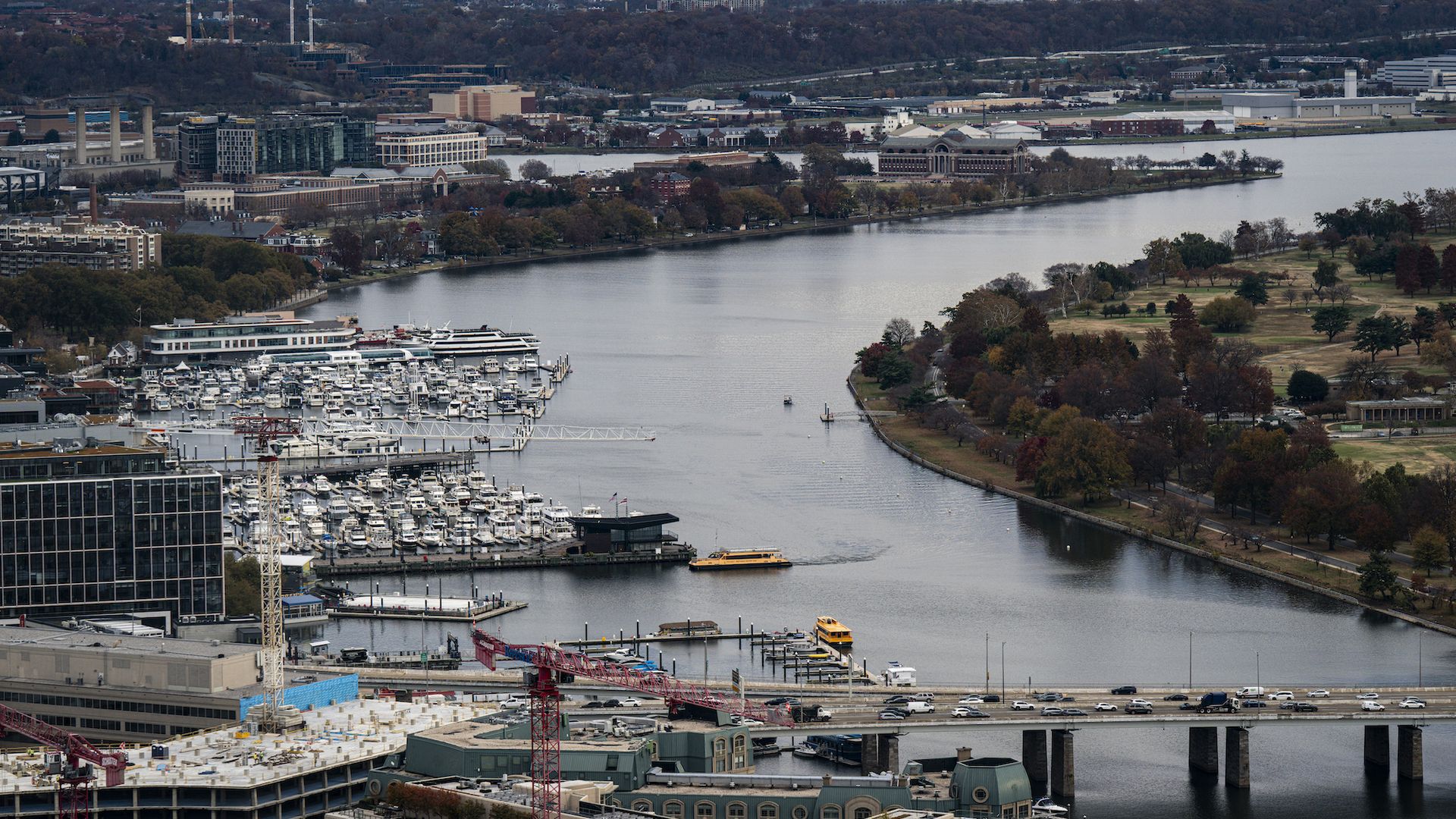 Aerial view of a the D.C. Wharf waterfront with a marina filled with boats, bridges with traffic, yellow water taxis on the river, and autumn trees lining the shore under a cloudy sky.