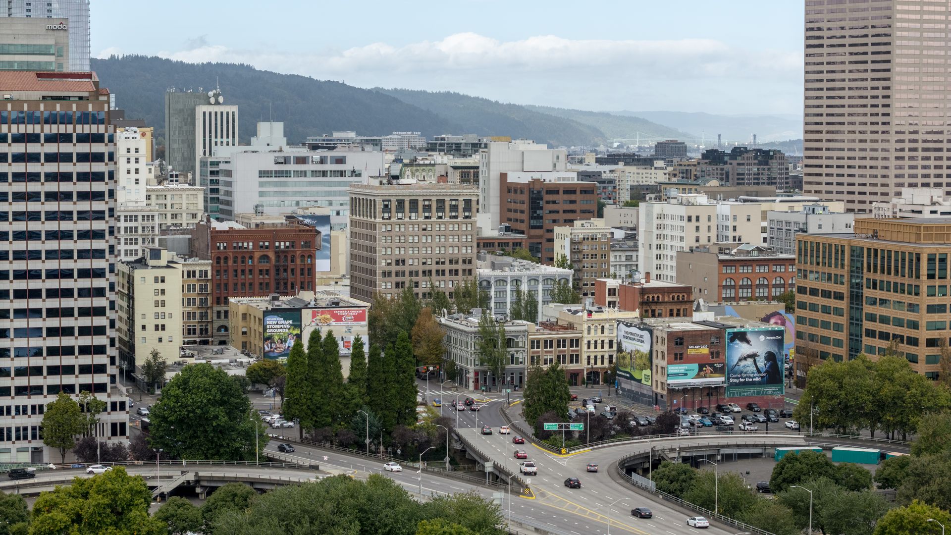 View of a cityscape with mid-rise buildings, a curved highway with cars, and green trees in the foreground under a partly cloudy sky and distant hills.