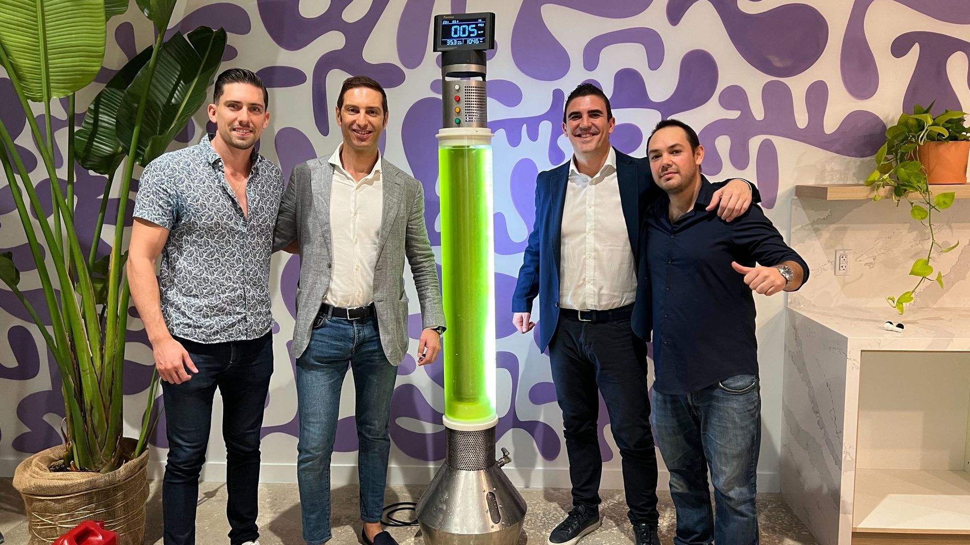 Four men stand beside a tall cylindrical machine with glowing green algae inside it and a digital display on top. 
