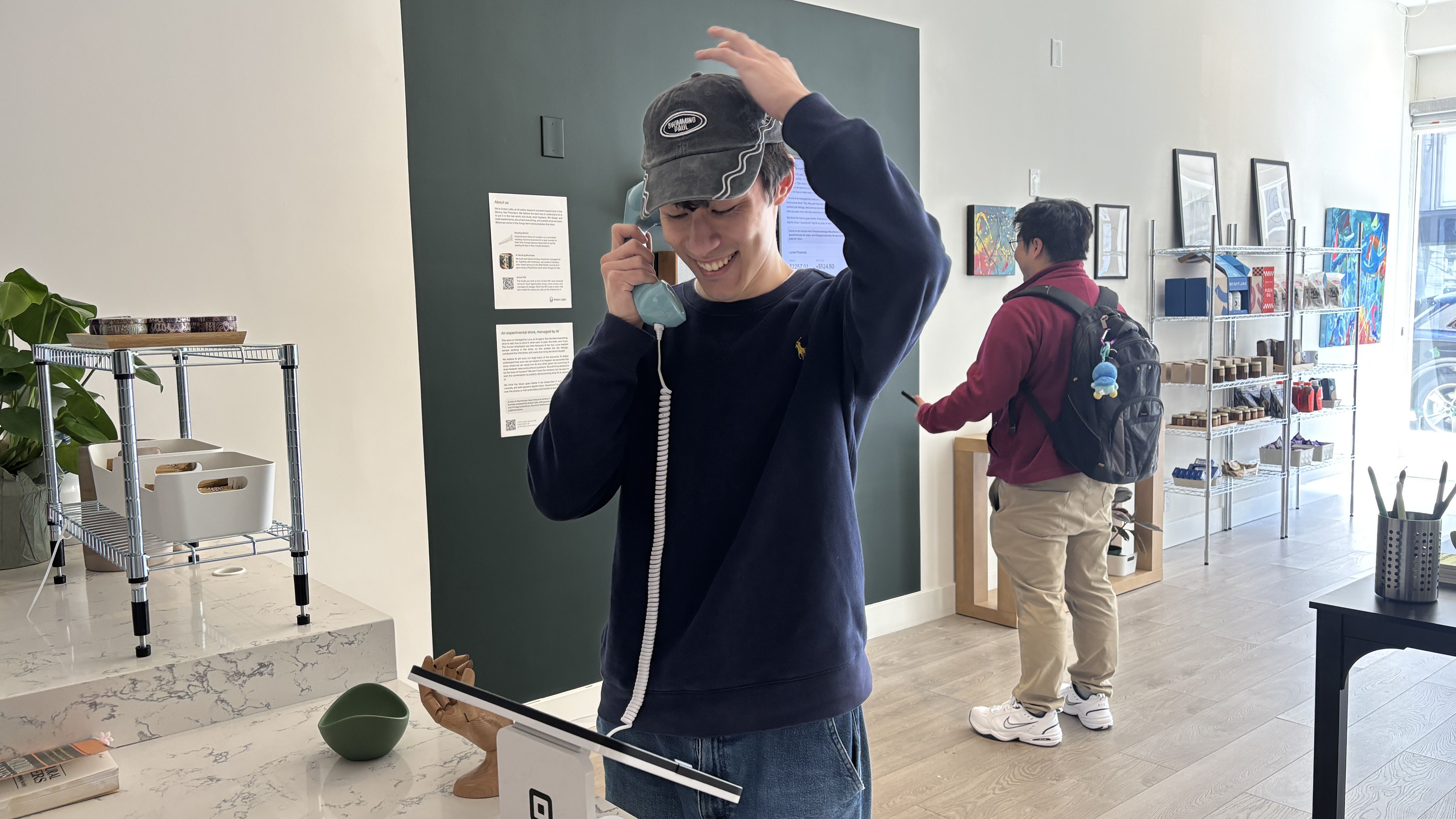 Inside a bright shop, a smiling young man in a navy sweater and cap talks on a vintage teal phone, adjusting his hat; another shopper with a backpack browses art and shelves in the background.