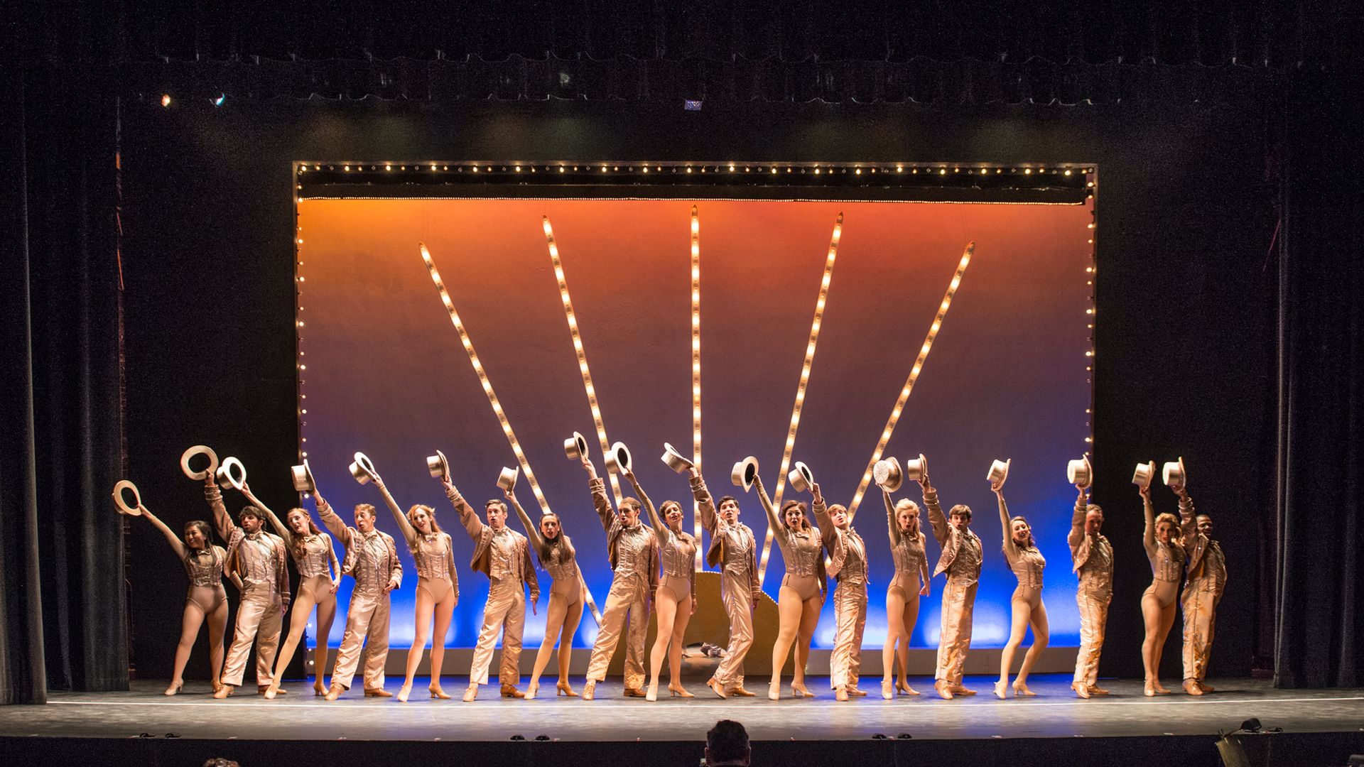 About two dozen performers, wearing gold, stand in a line with hats held in the air.