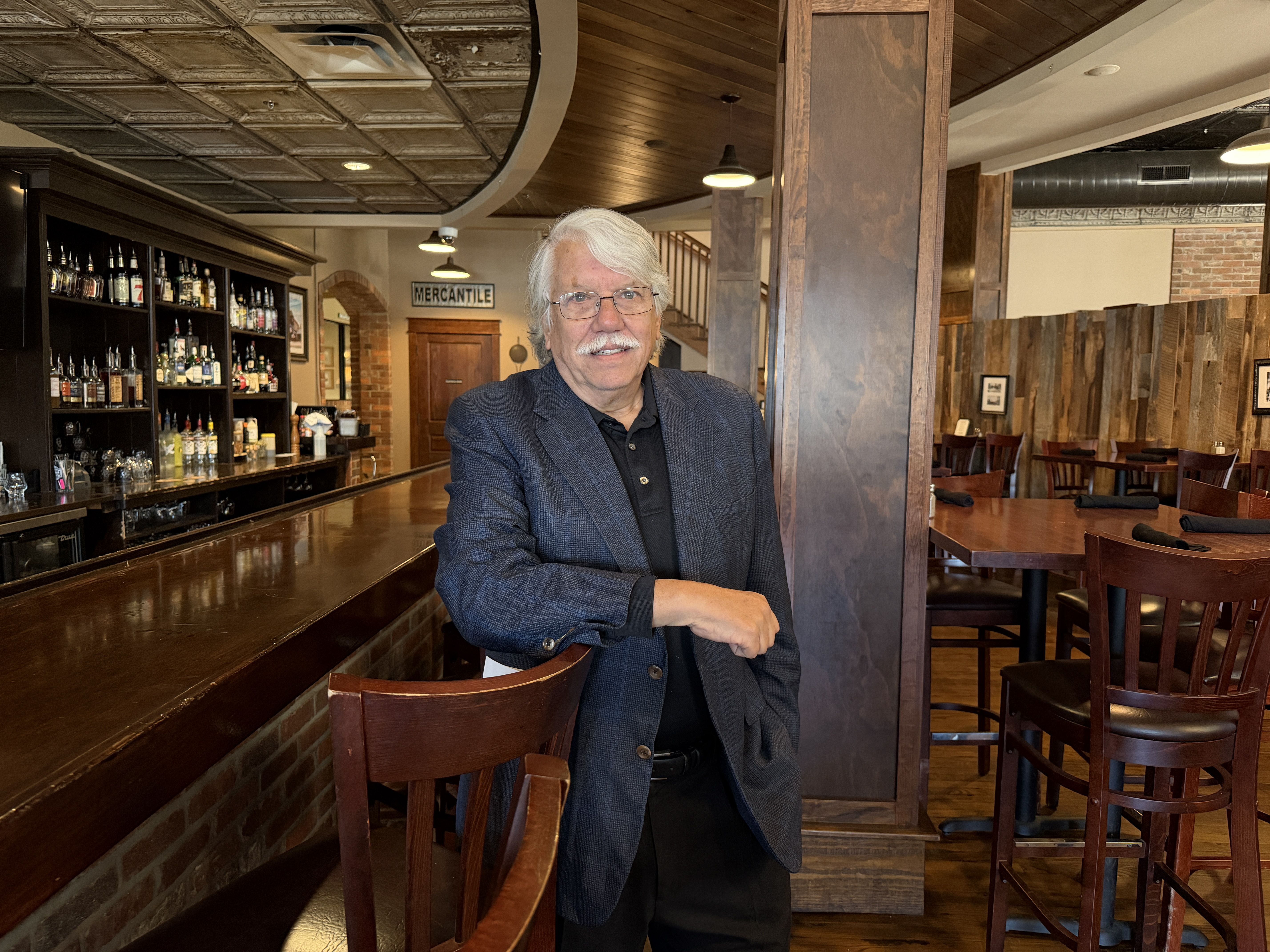 Older man with white hair and mustache wearing glasses, navy blazer, and black shirt standing in a wood-accented bar with shelves of liquor bottles and empty wooden tables.