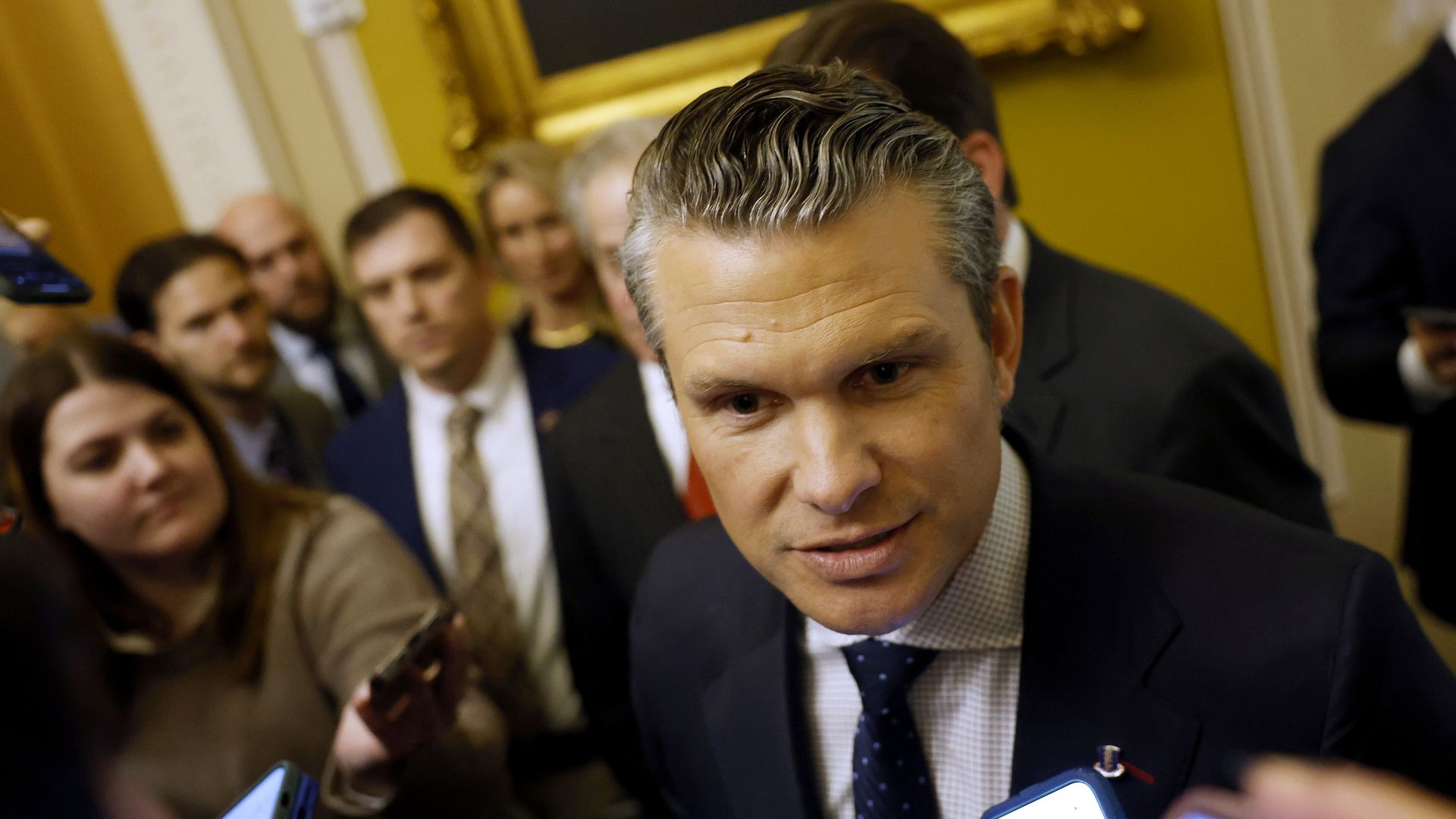 Pete Hegseth leans forward to answer a question while surrounded by reporters.