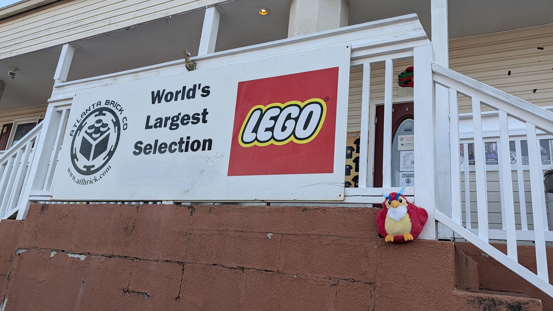 White sign with black text "World's Largest Selection" and red LEGO logo on building with white railing, brick wall, and a colorful red, blue, and yellow plush toy on the ledge.