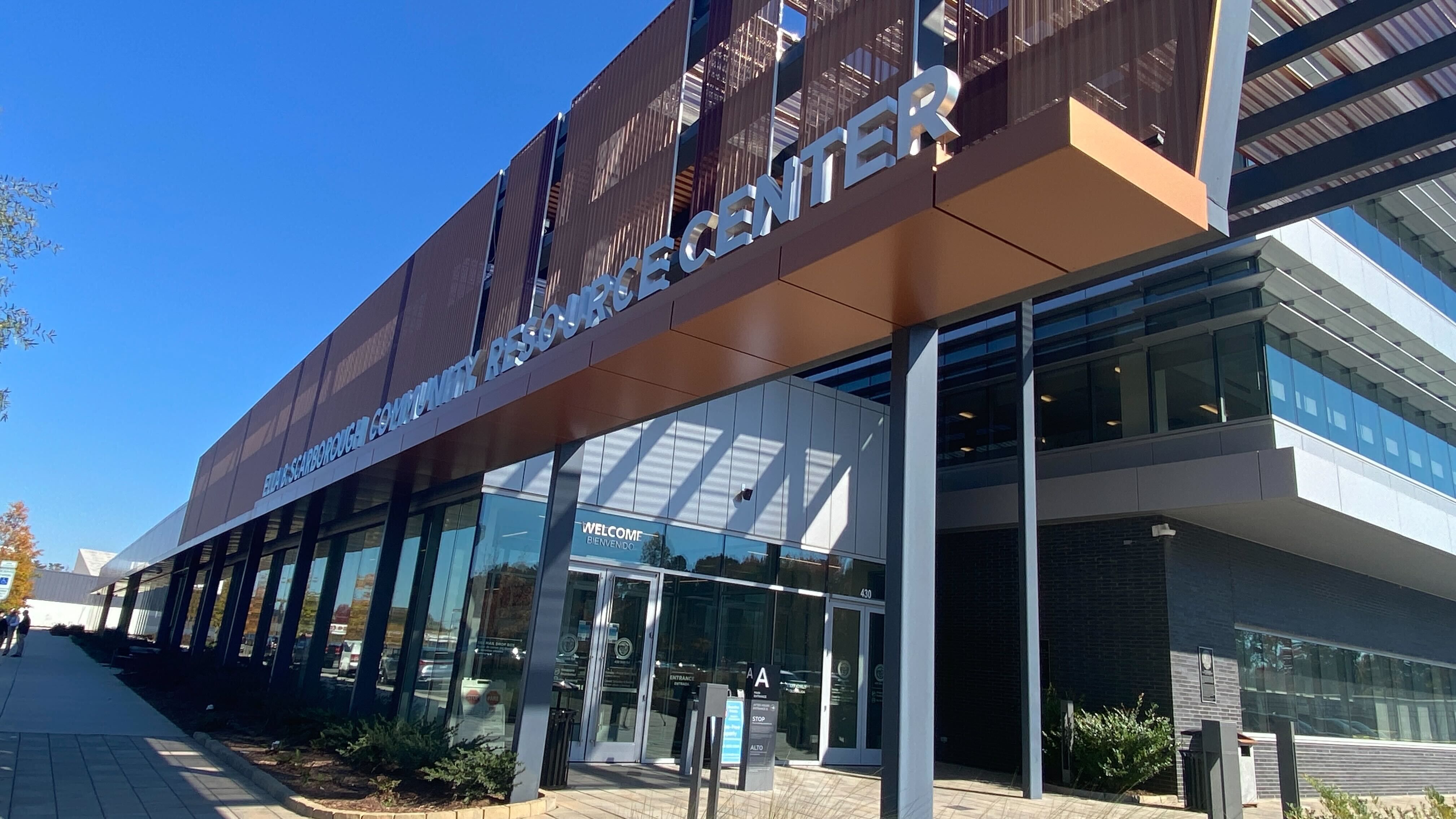 Modern building with glass doors and large windows under blue sky, labeled ELLA SCARBOROUGH COMMUNITY RESOURCE CENTER in white letters above entrance.
