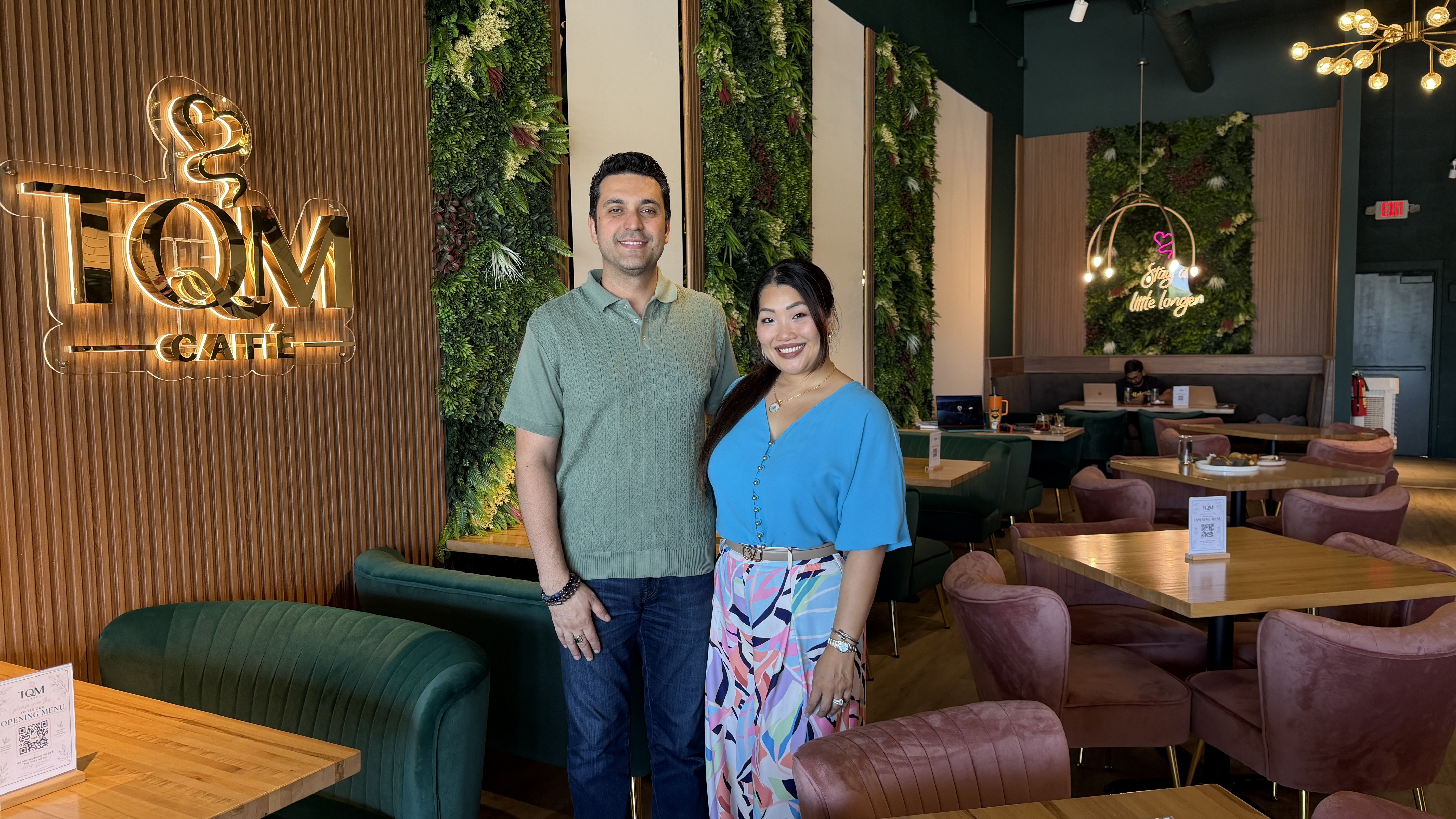 Two smiling people stand in a stylish cafe with wood panels and lush green plant walls. A glowing "TQM CAFE" sign sits at left, while a neon "Stay a little longer" sign glows in the background.