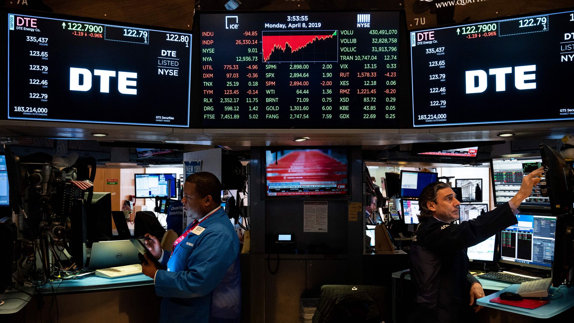 In this image, two men stand facing away from each other on the stock market trading floor, with three screens above and behind them.