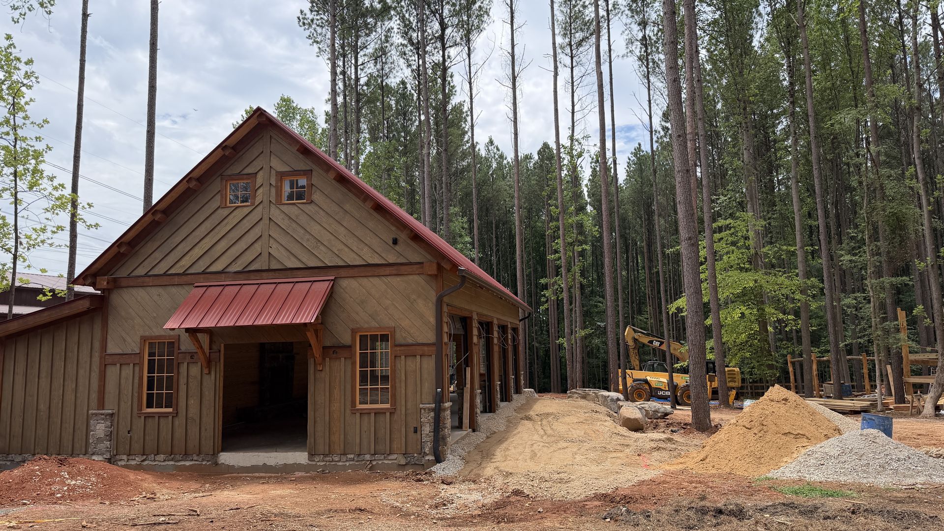 A wooden cabin with a red slanted roof sits at a construction site, surrounded by tall pine trees. Dirt piles and yellow excavators are visible in the background.