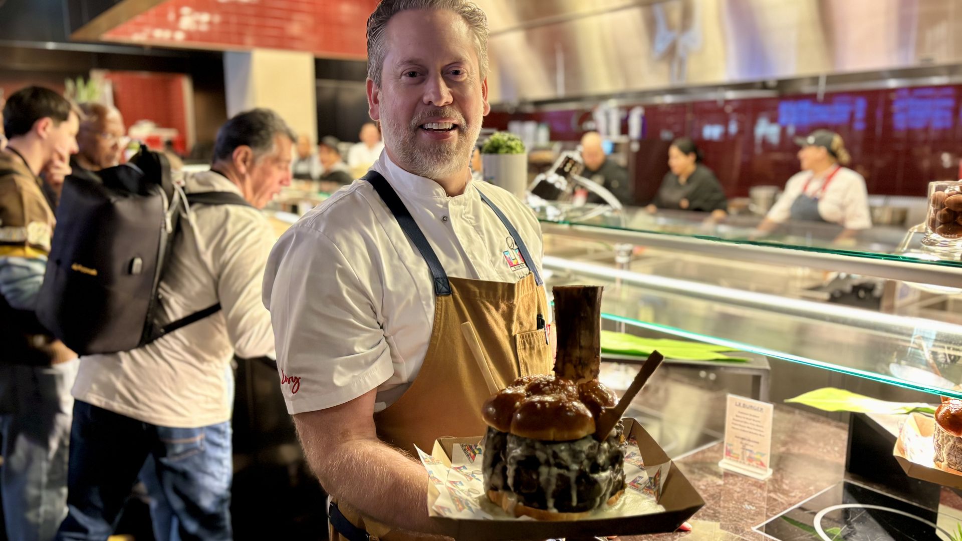 Smiling chef in a white shirt and tan apron holding a large, stacked burger with a pretzel bun and bone, standing behind a glass counter in a busy kitchen with people in the background.