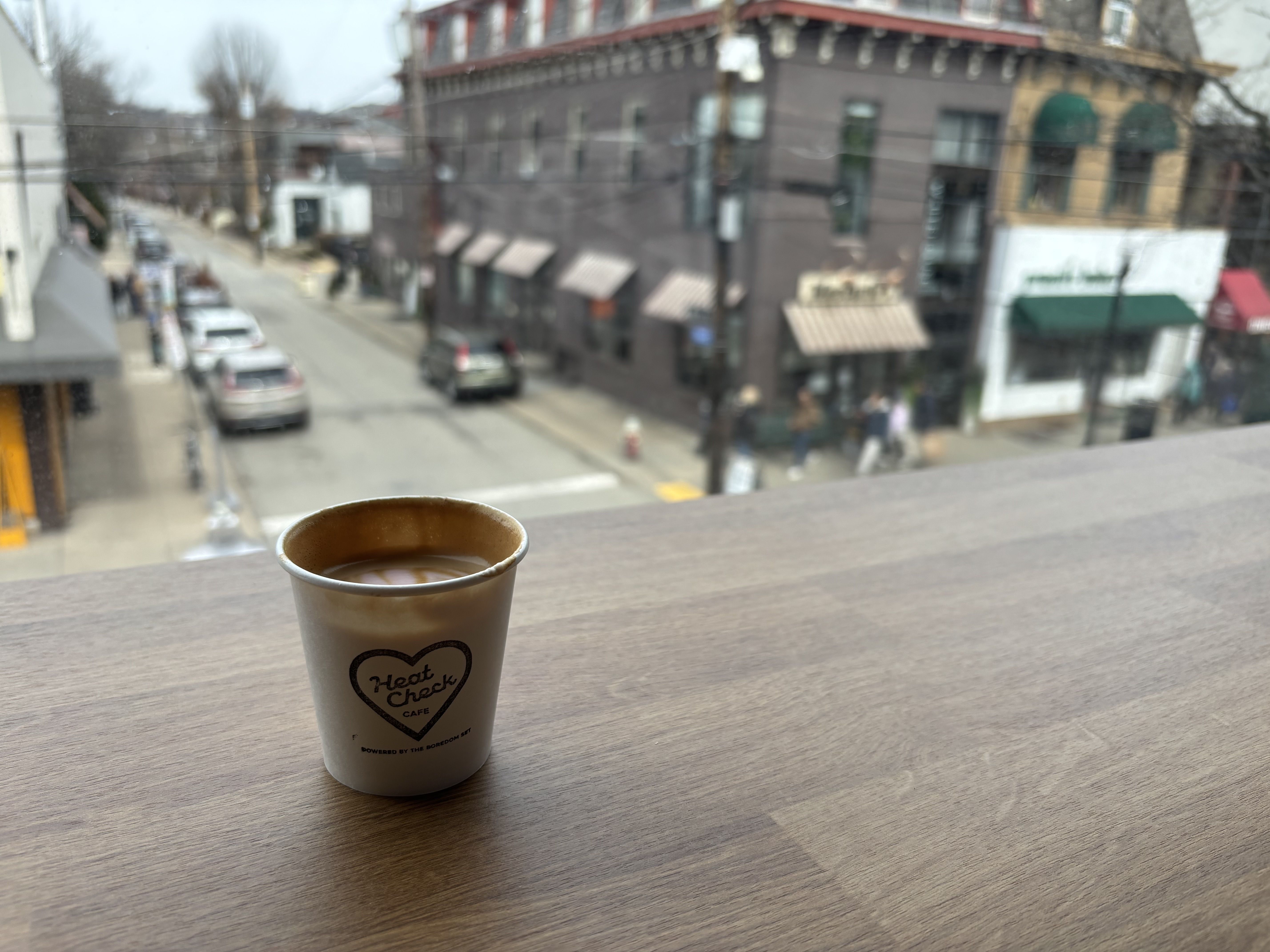 Paper cup with coffee on wooden table inside cafe, window view of street with cars, pedestrians, and buildings with awnings on cloudy day.
