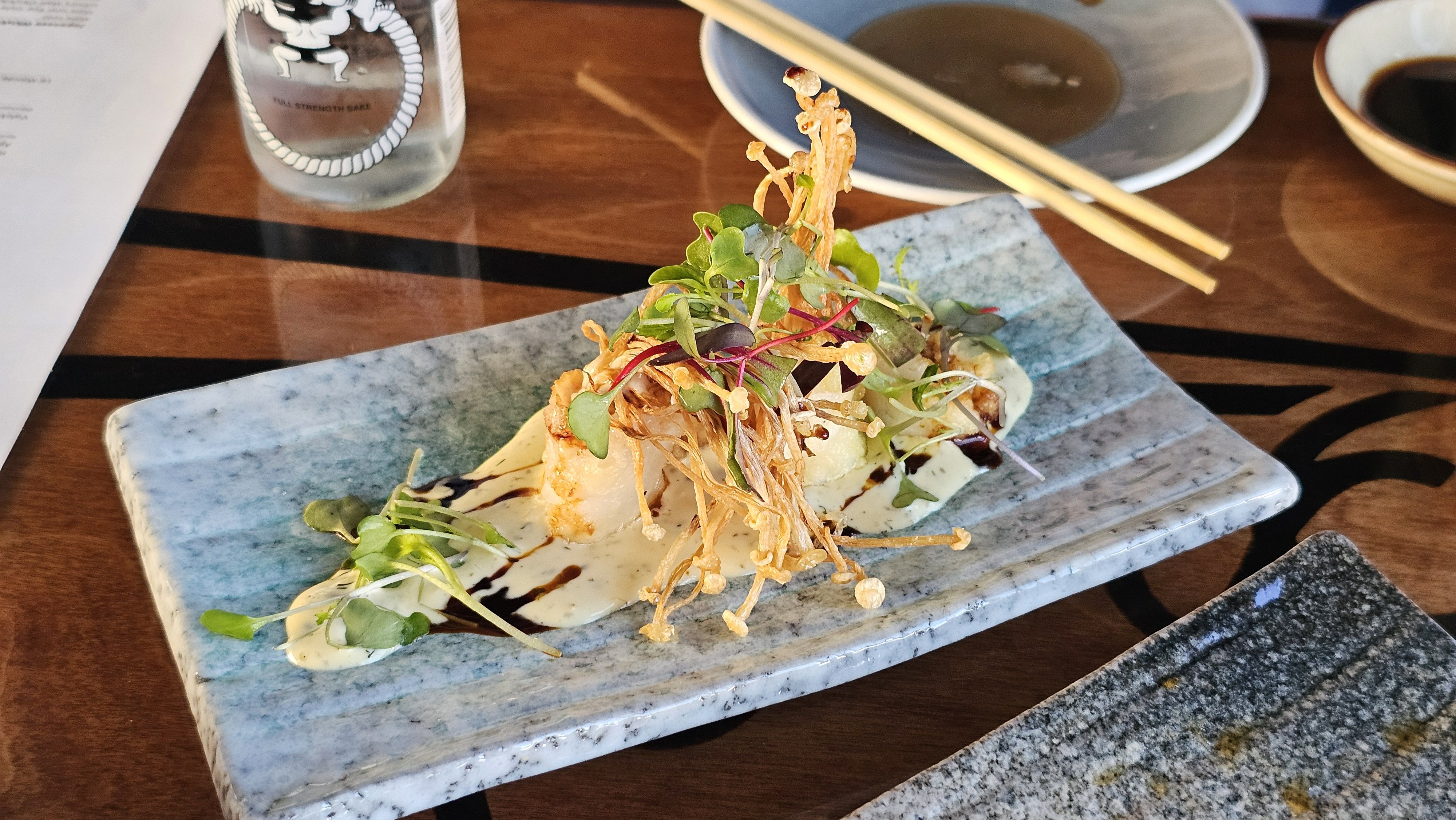 Plated dish with crispy fried strands and microgreens atop pale sauce on a blue-gray rectangular plate; chopsticks and a bottle appear on a wooden table in the background.