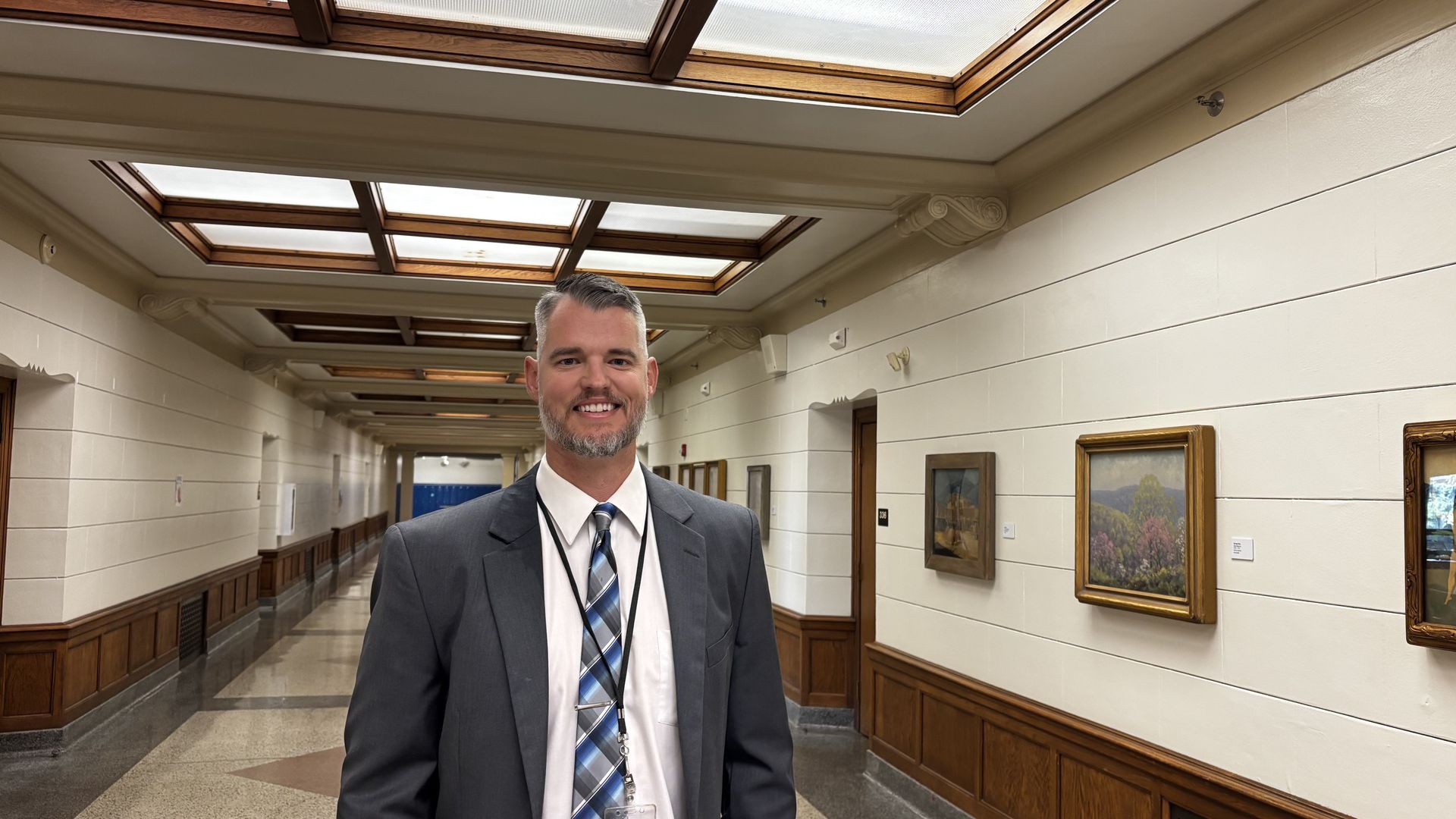 A smiling man in a gray suit and striped tie stands in a long hallway with wood-paneled walls and framed paintings. Skylight panels on the ceiling let in soft natural light.