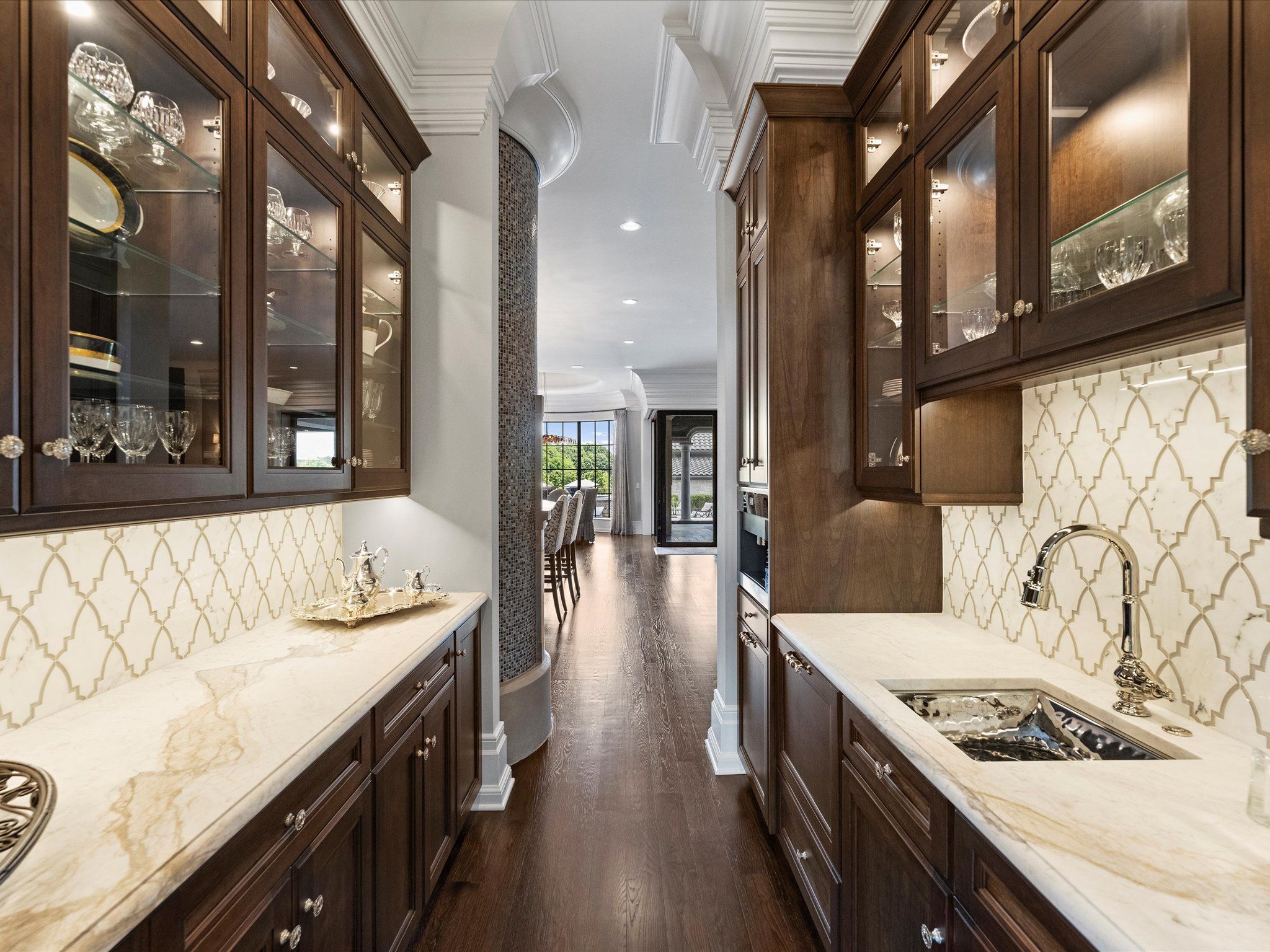 Narrow kitchen with dark wood cabinets, marble countertops, glass-front upper cabinets displaying glassware, white patterned backsplash, and a view through to a dining area with tall windows.