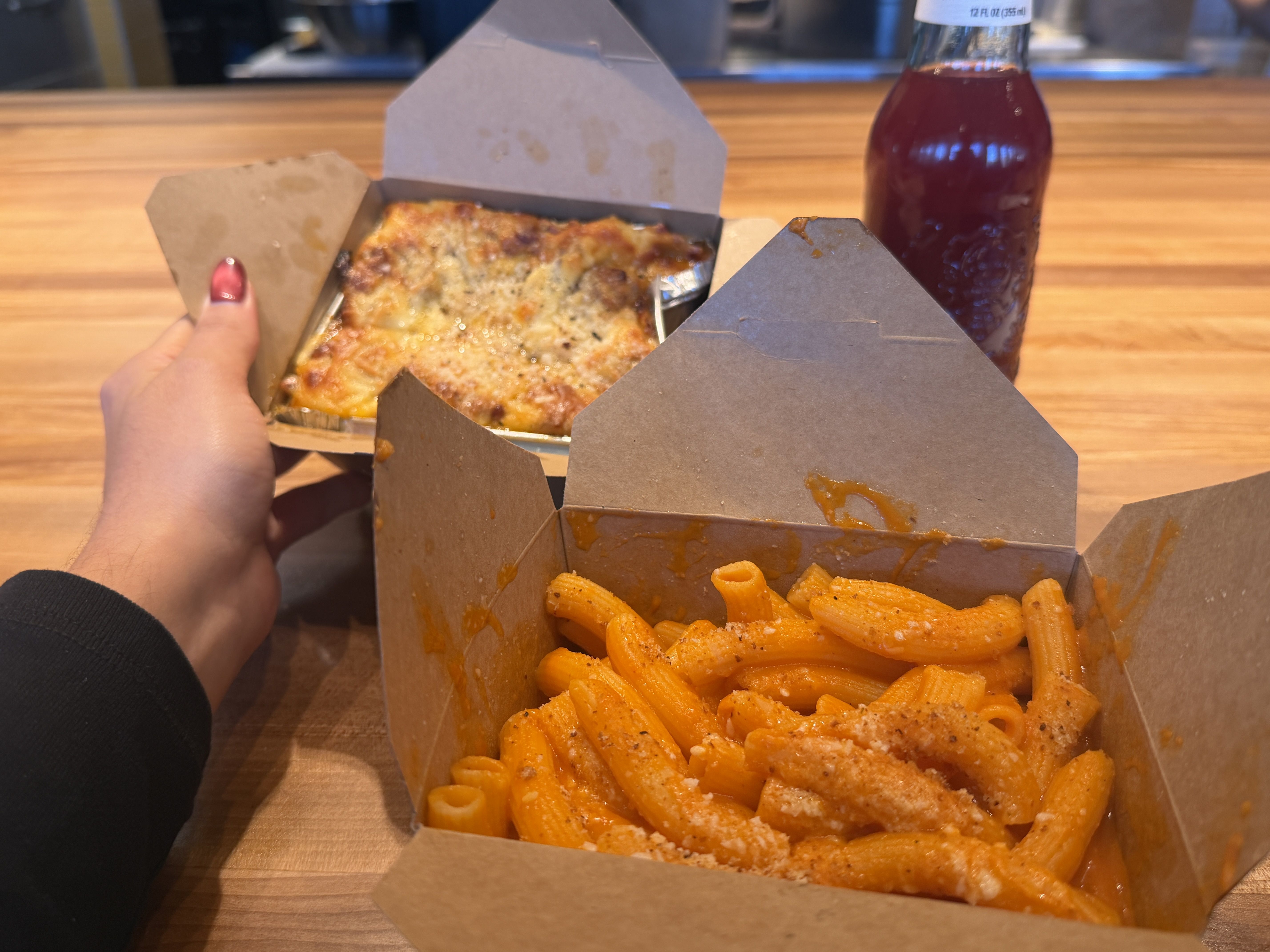 Hand holding a takeout box with lasagna, another takeout box of baked ziti with red sauce and cheese on a wooden table, and a bottle of dark red beverage in the background.